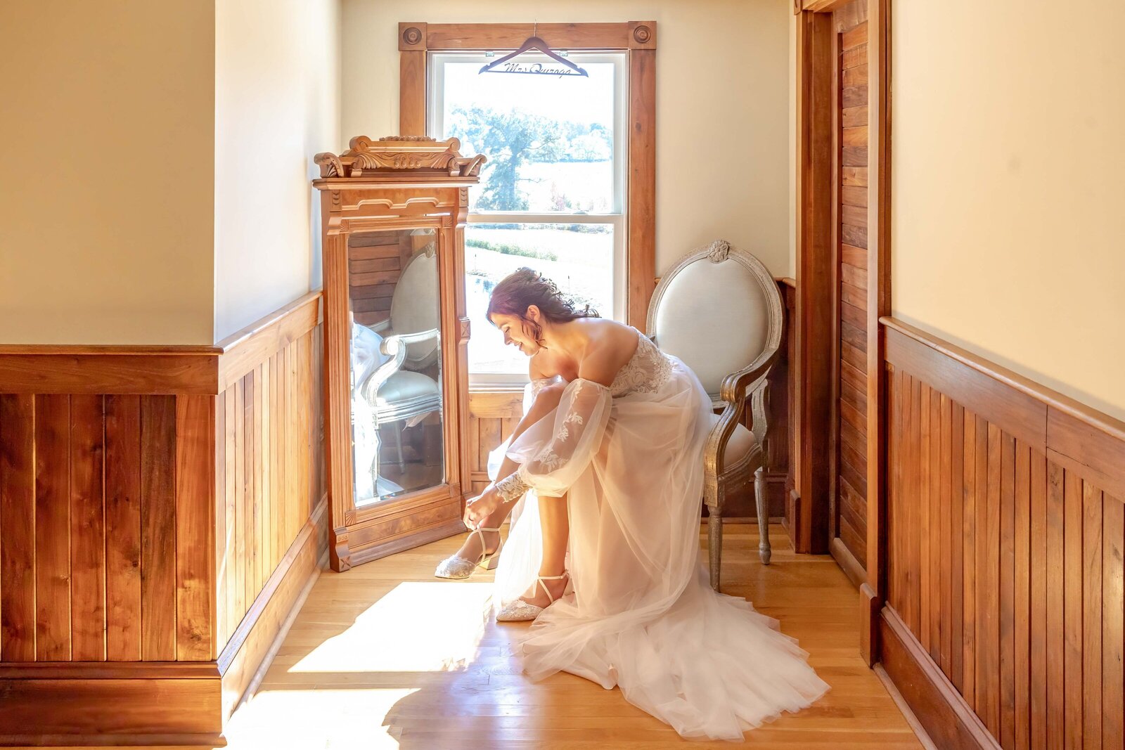 A bride in a flowing gown adjusts her shoes in a rustic bridal suite with wooden walls and vintage decor, captured during a Apex, NC wedding, showcasing elegant and authentic preparation moments.