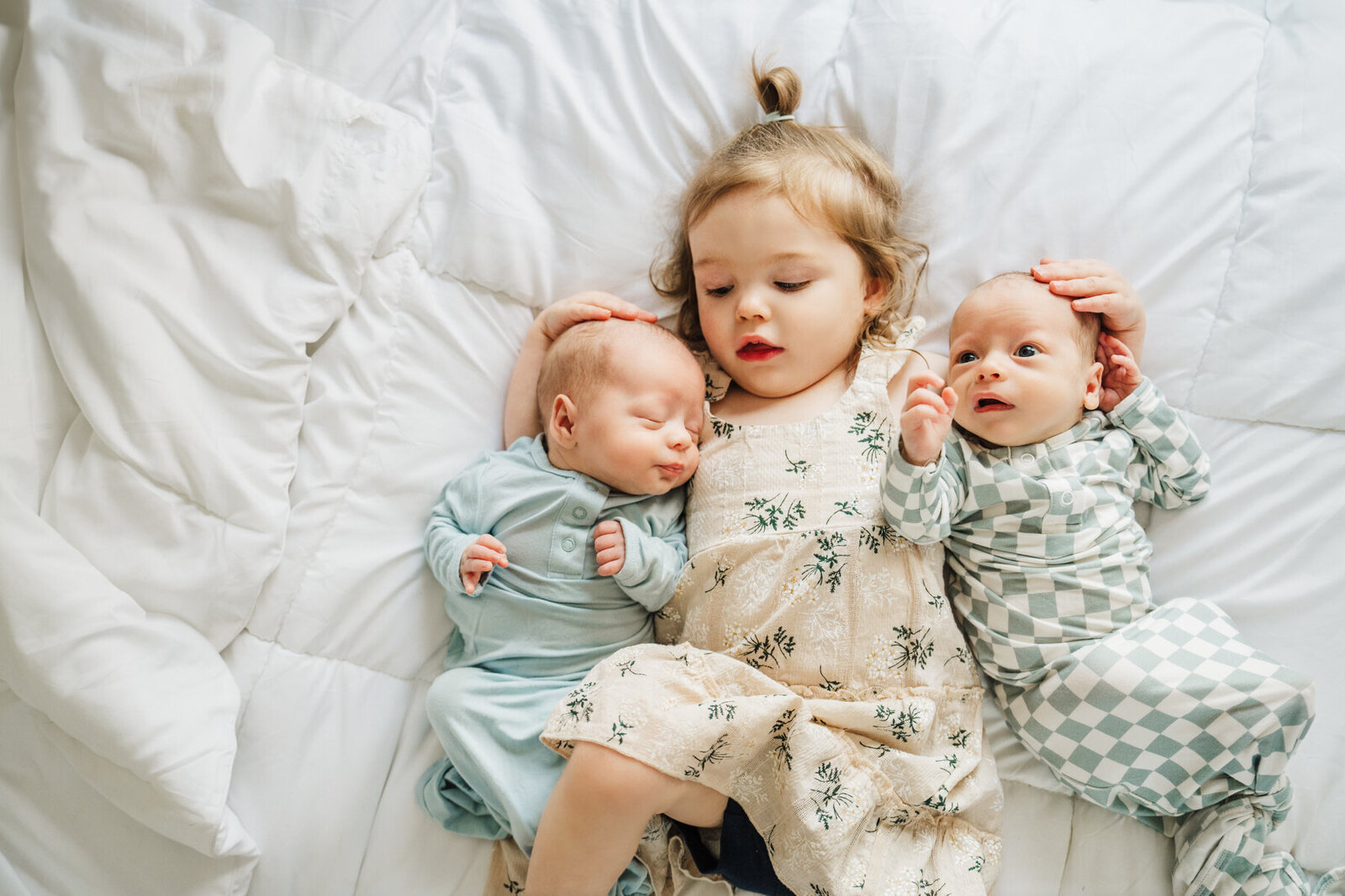 big sister snuggles twin brothers while laying on white bedspread