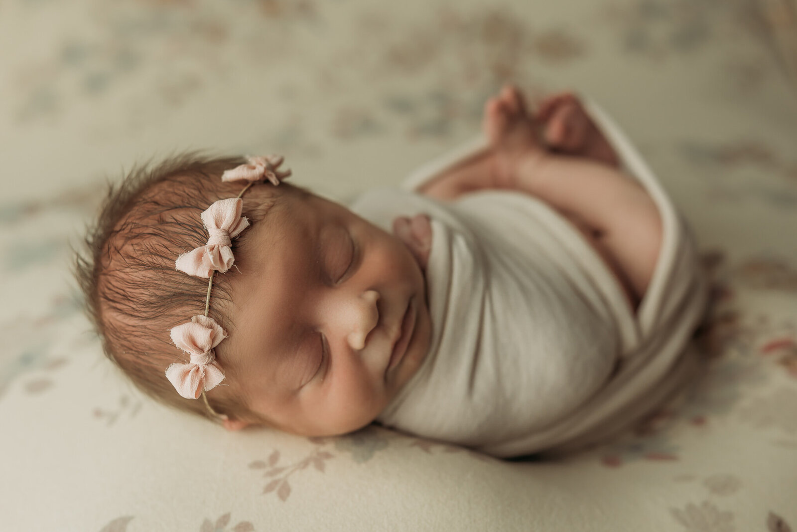 Baby girl swaddled in a tan swaddle with a headband of tiny pink bows sleeping during her newborn photos