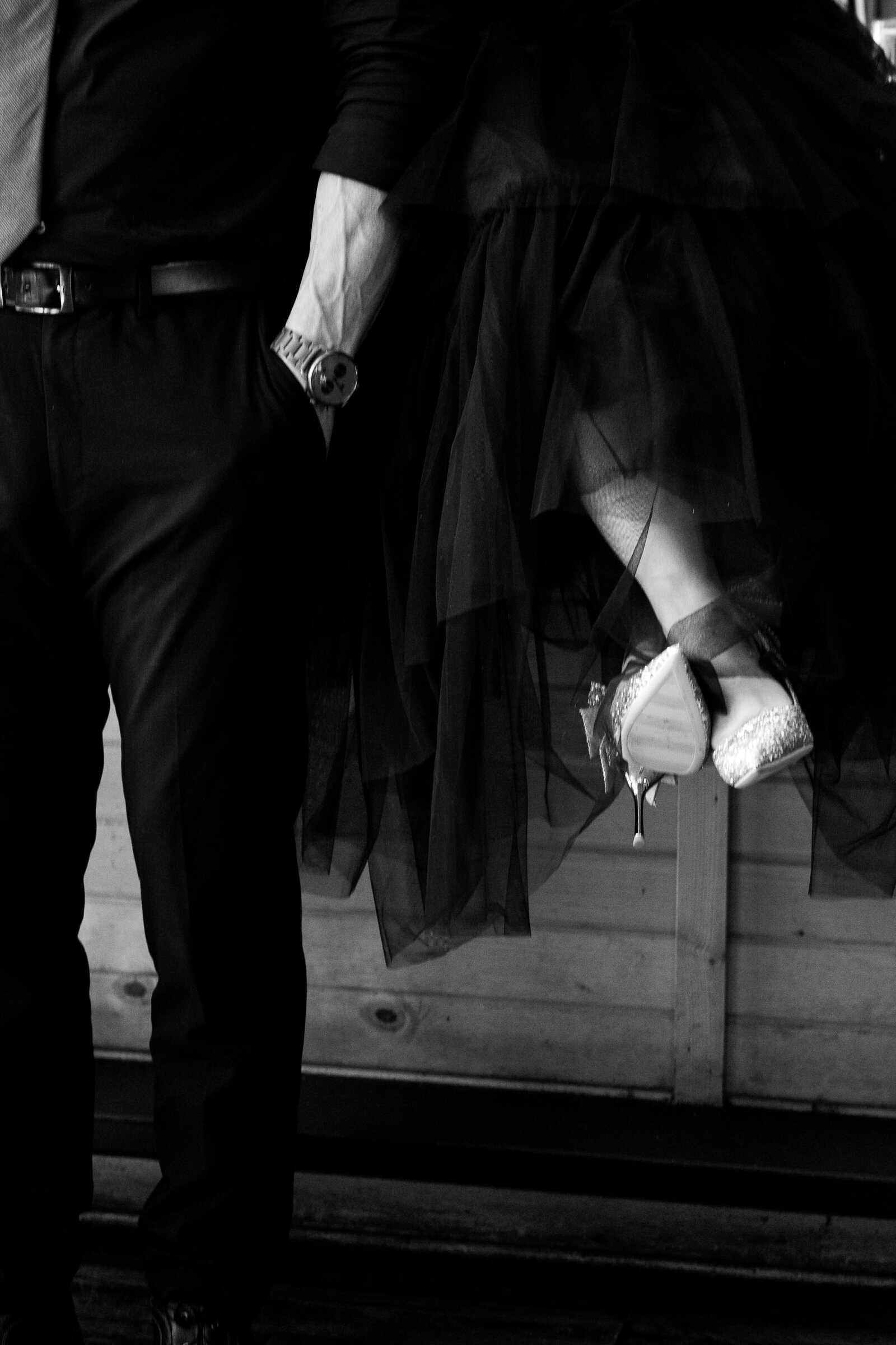 Cinematic engagement photo of a couple sitting on the bar. She has heels and he has a watch on. They are both wearing black.