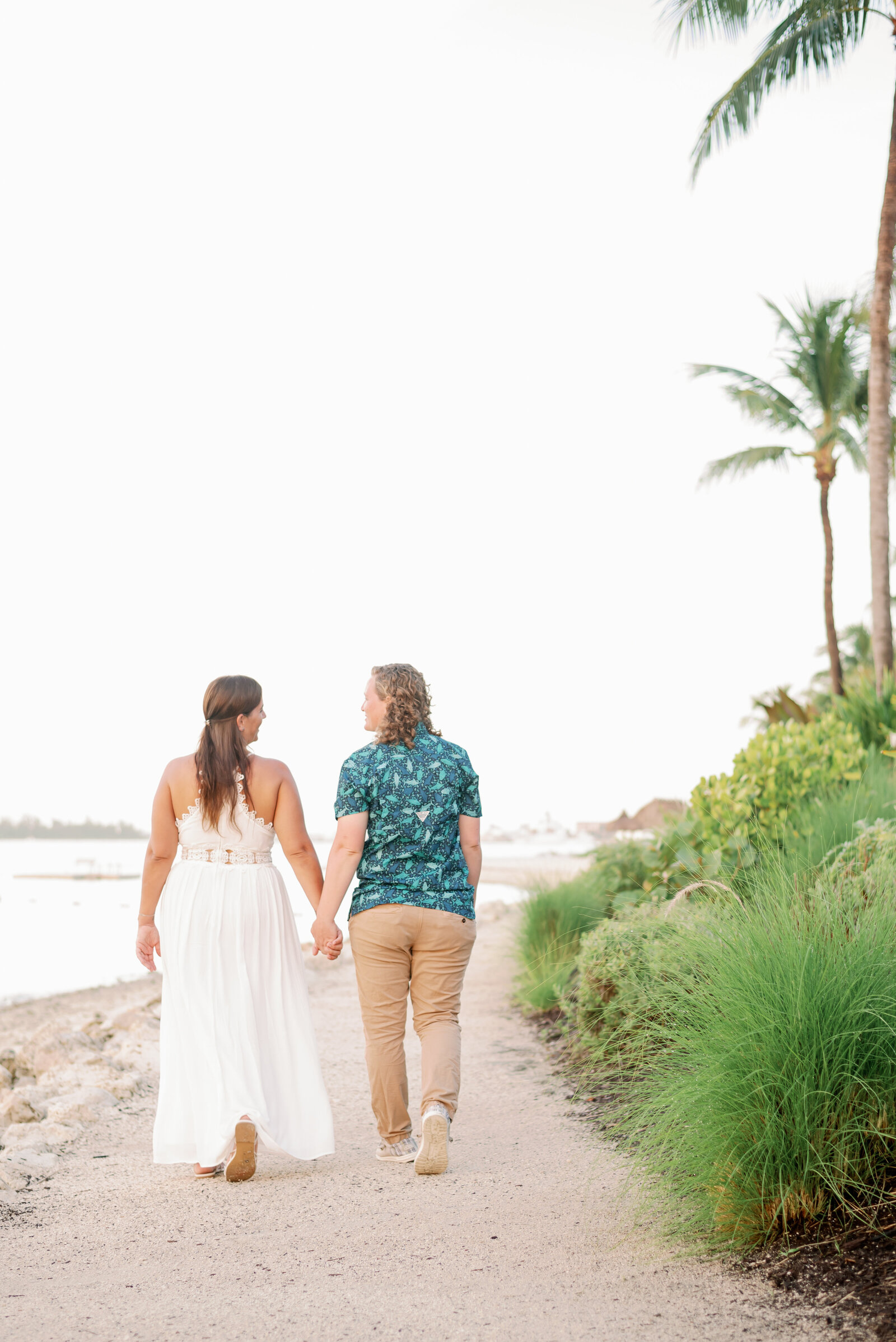 LGBTQ+ engaged couple holding hands and walking away from the camera on a beach while looking at each other