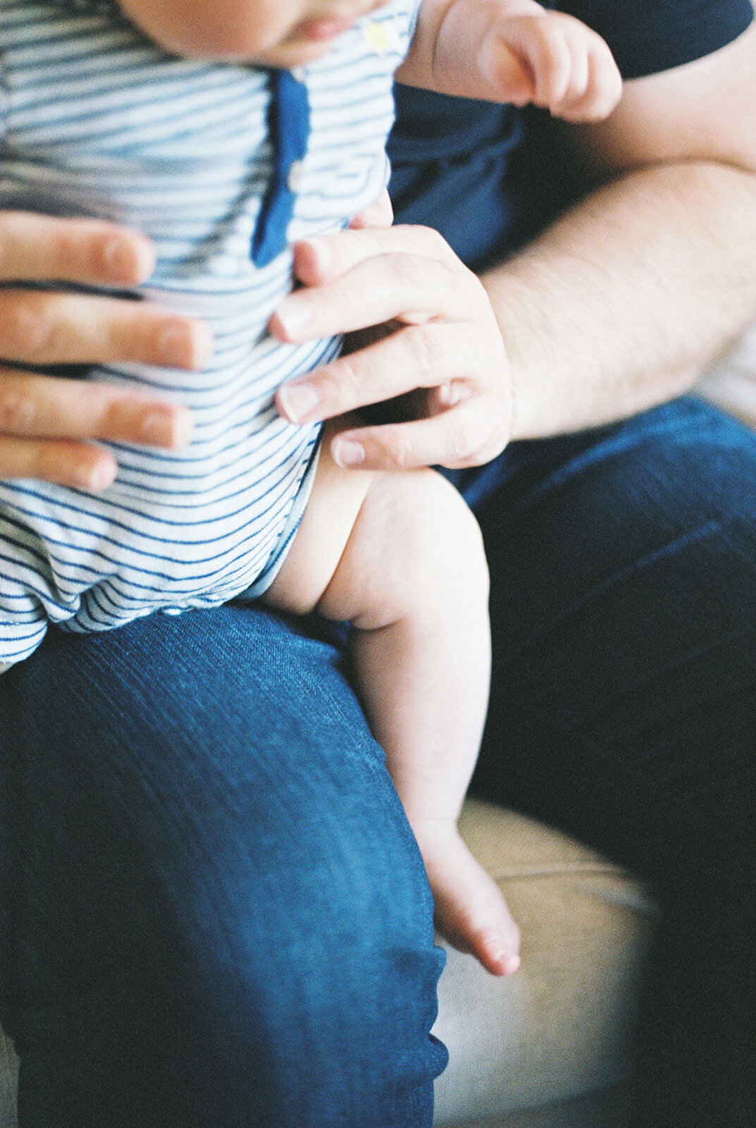 Close-up of a father holding a baby in striped onesie, capturing the baby’s tiny hands and chubby legs.