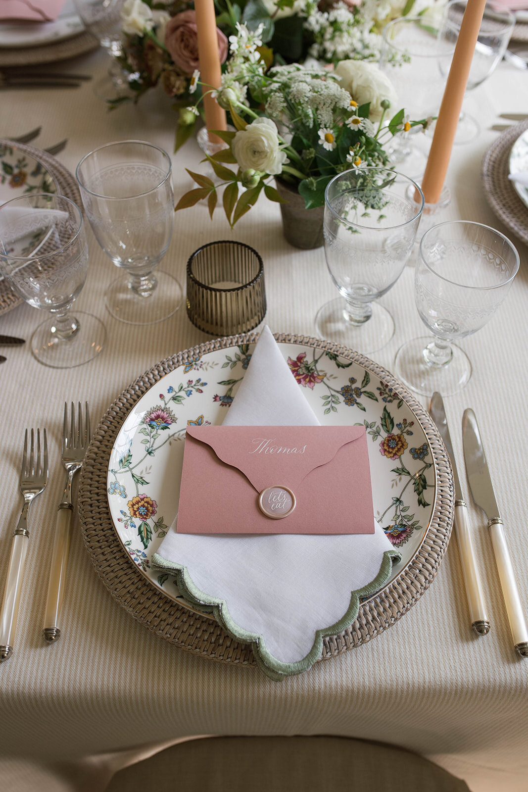 The reception table setting with neutral cream linen, a wicker charger, and floral china plate embellished with a napkin and custom pink place card.
