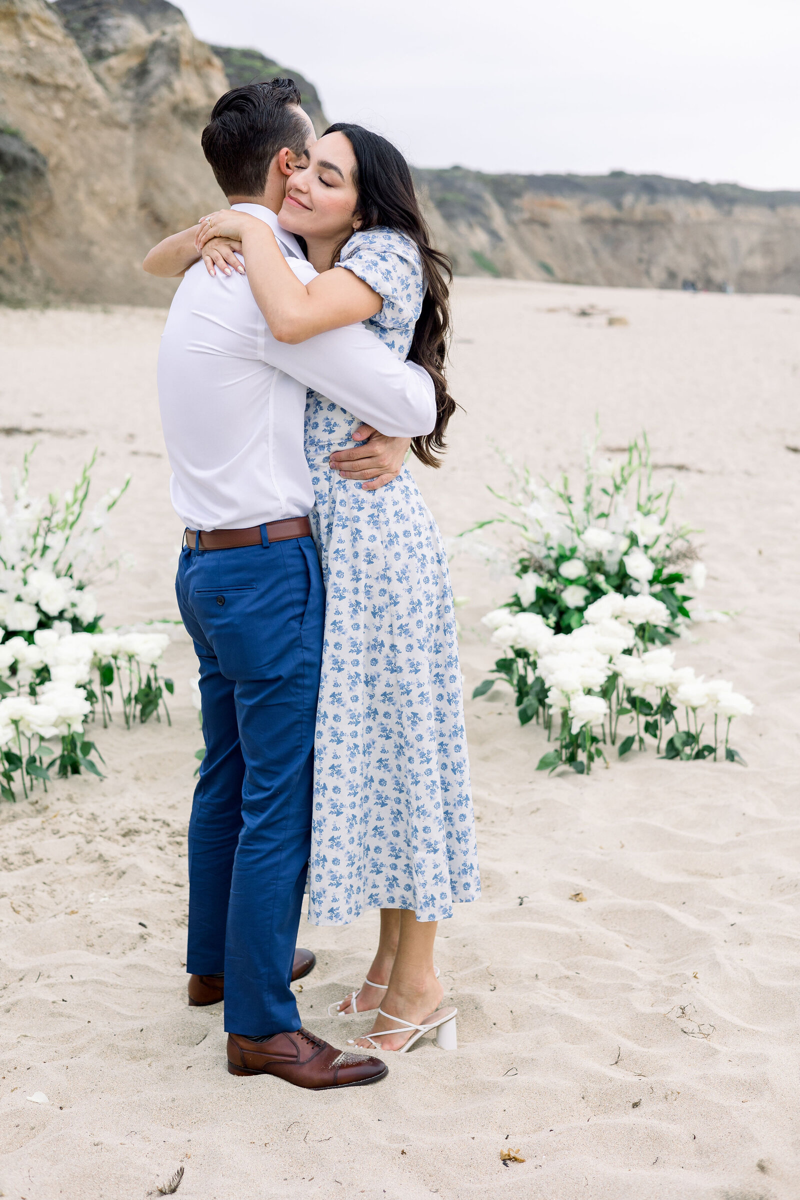 beach engagement photos for a surprise proposal on the beach with man and woman hugging at each other with the cliff side in the distance captured by wedding photographer bay area