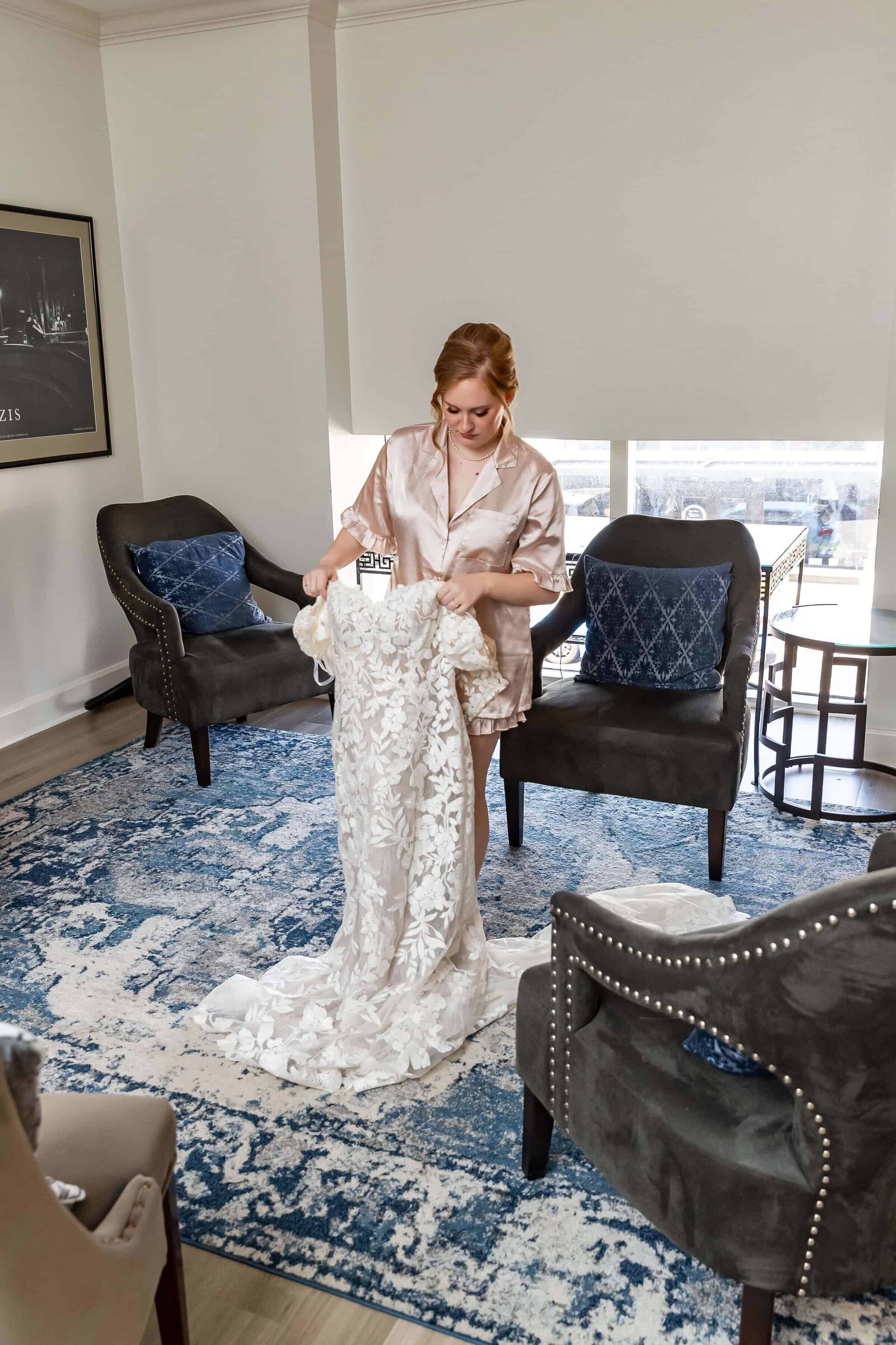 A woman in a satin robe stands in a bright room, holding up a long, white lace wedding dress over a blue patterned rug, surrounded by gray chairs with decorative pillows.