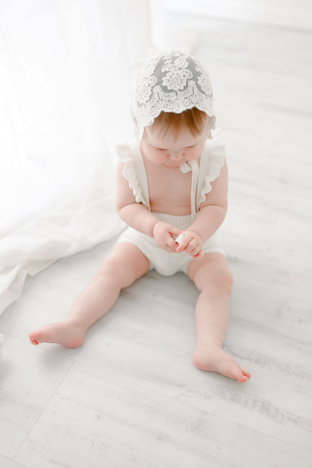 Baby mit einer weißen Rüschenhaube spielend auf dem Boden bei einem Babyshooting bei Bielefeld, Paderborn, Gütersloh und Verl.