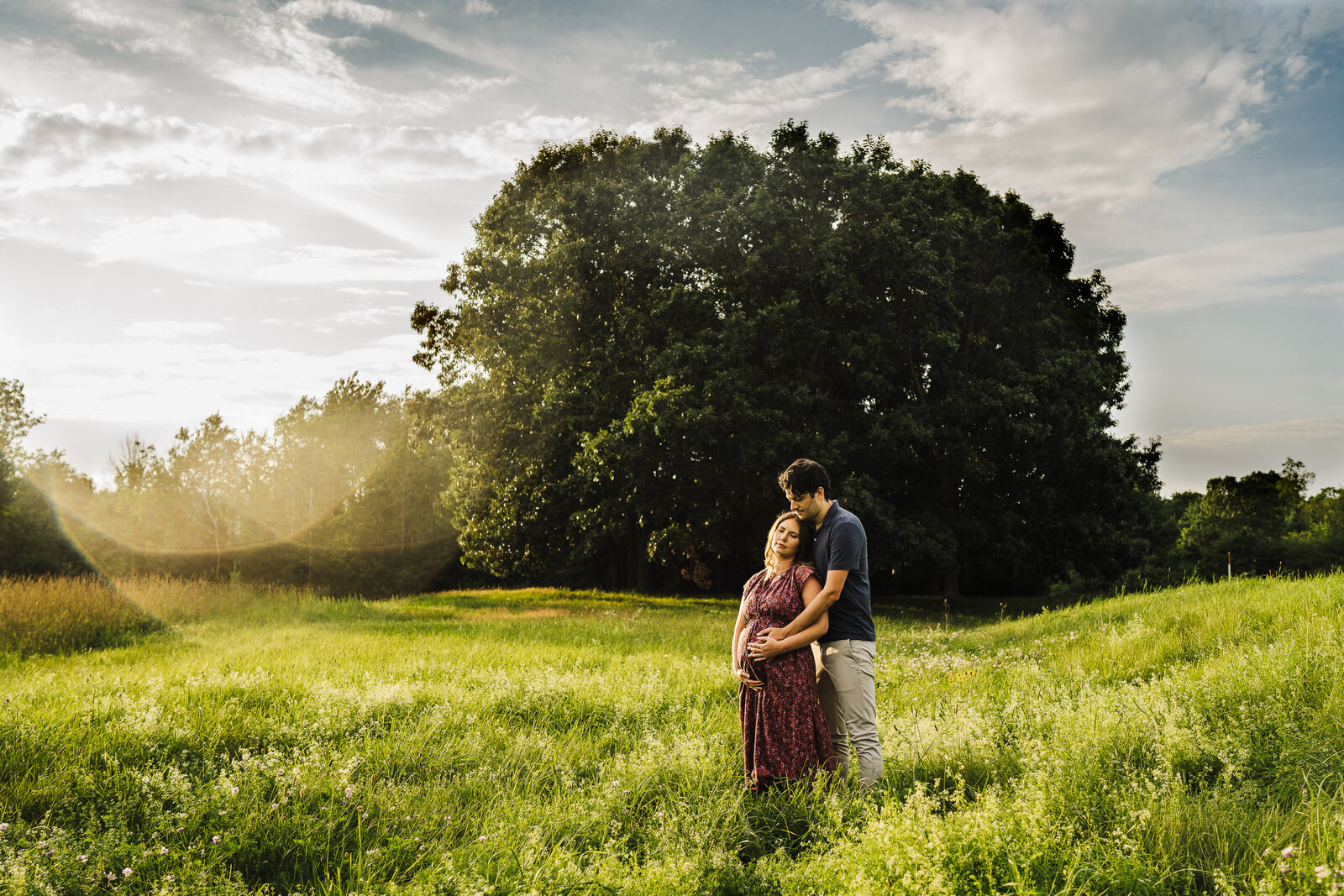 pregnant couple stands in field at sunset