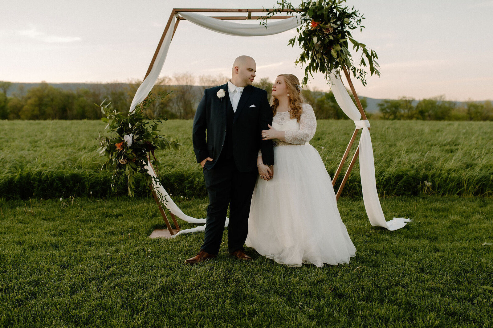 couple standing under hexagon wooden wedding arbor with white fabric, florals, and greenery