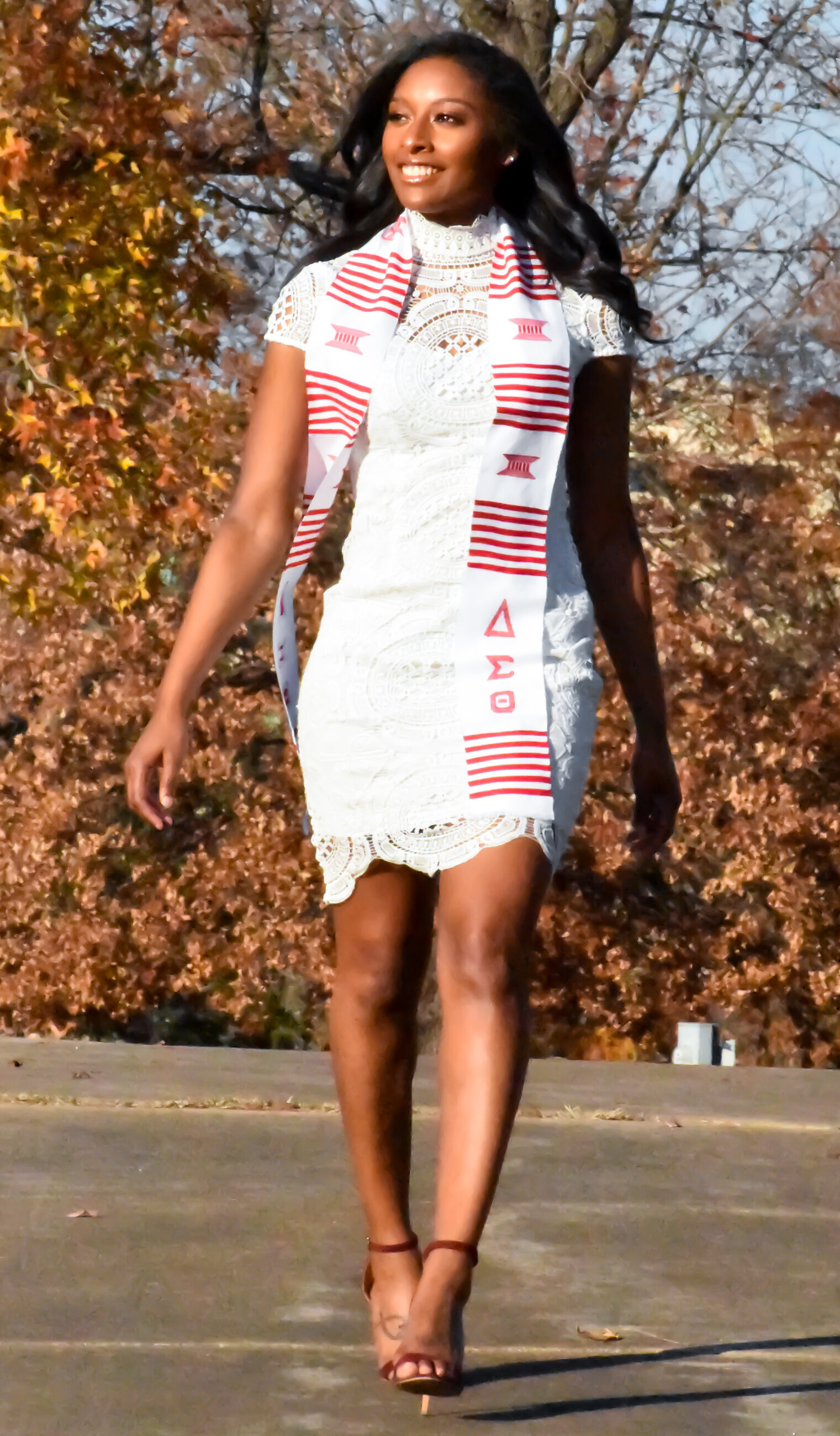 a senior dressed in a white dress with her red and white stole draped over her shoulder sehs walking and smiling. photographed by Millz Photography in Greenville, SC