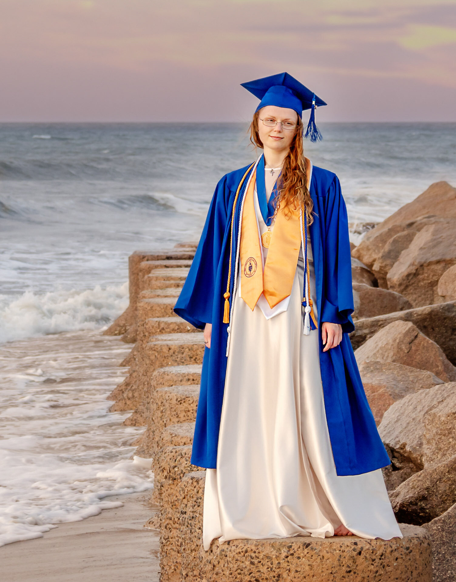 A Cape Fear Community College senior in a blue cap and gown standing on rocks by the ocean in North Carolina. This striking photo captures the achievement of graduation, ideal for those seeking professional and scenic senior photography.