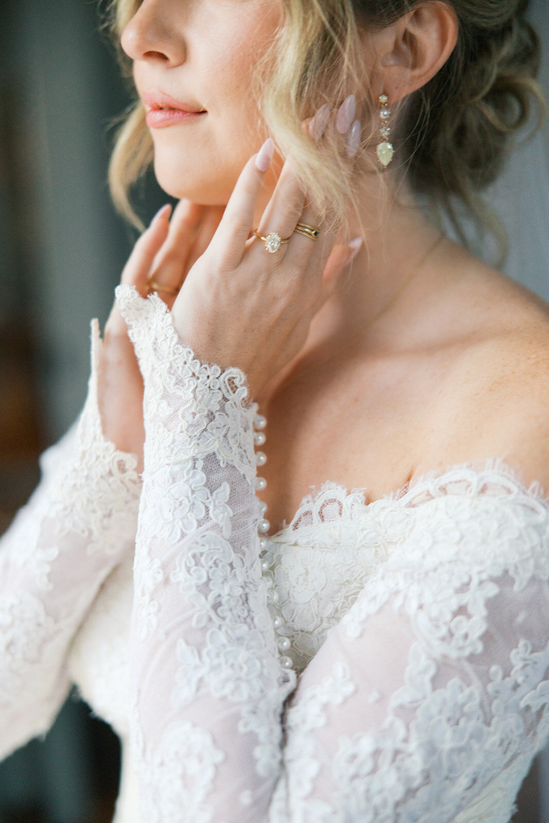 Bride with lace sleeves, off the shoulder wedding gown touches her cheeks with her oval engagement ring