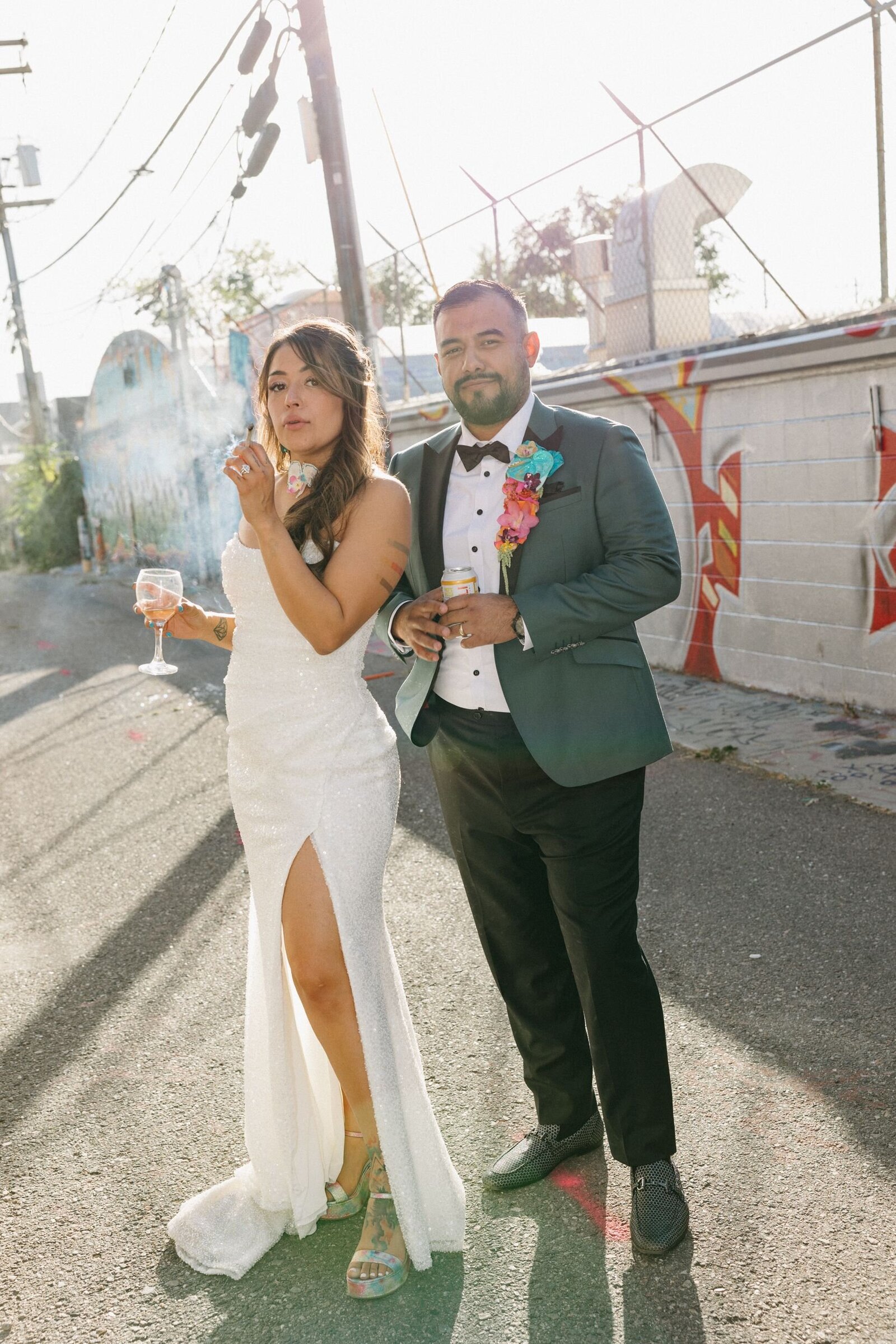 A bride and groom stand in a city alley holding drinks and smoking a joint