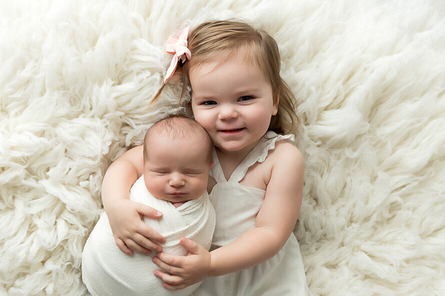Toddler girl holds her new baby sister on a fluffy white backdrop.