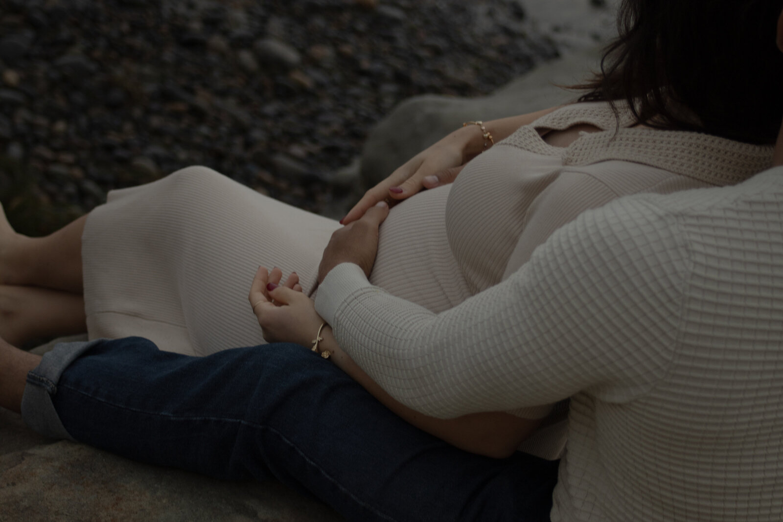 expecting parents sitting on cliff at oceanside while husband caresses baby bump