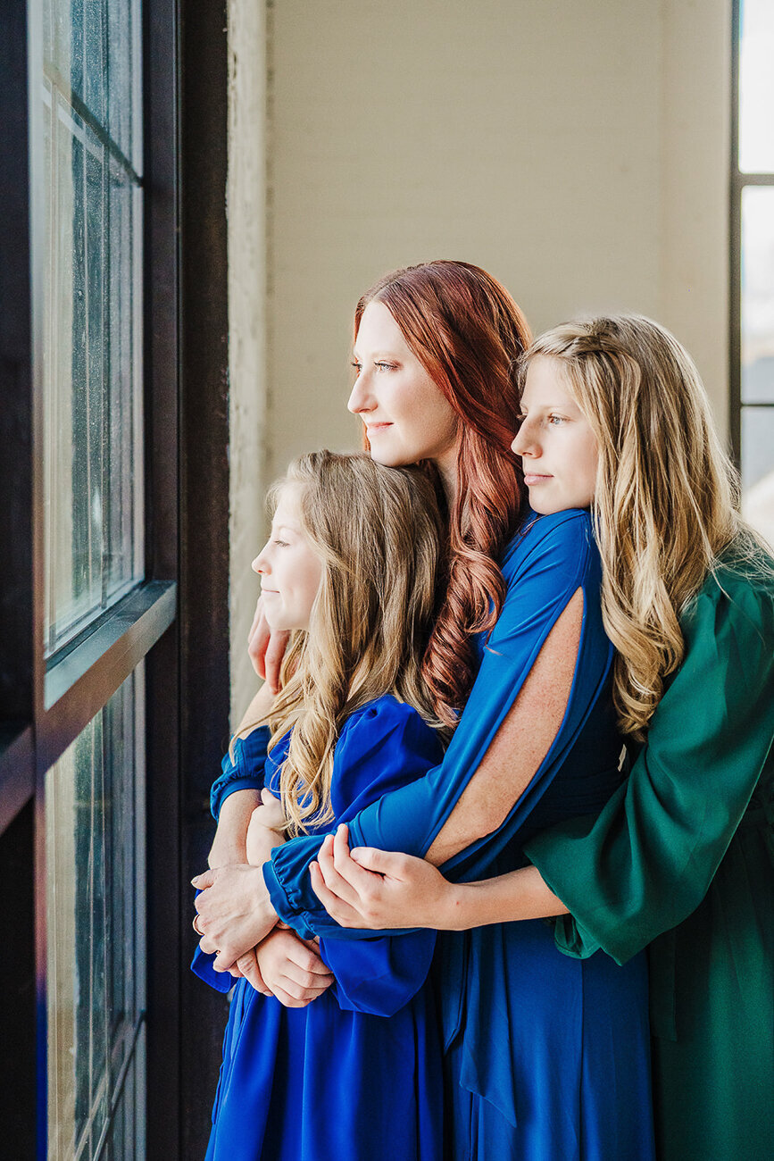 mother holds two daughters in jewel dresses close while looking out window