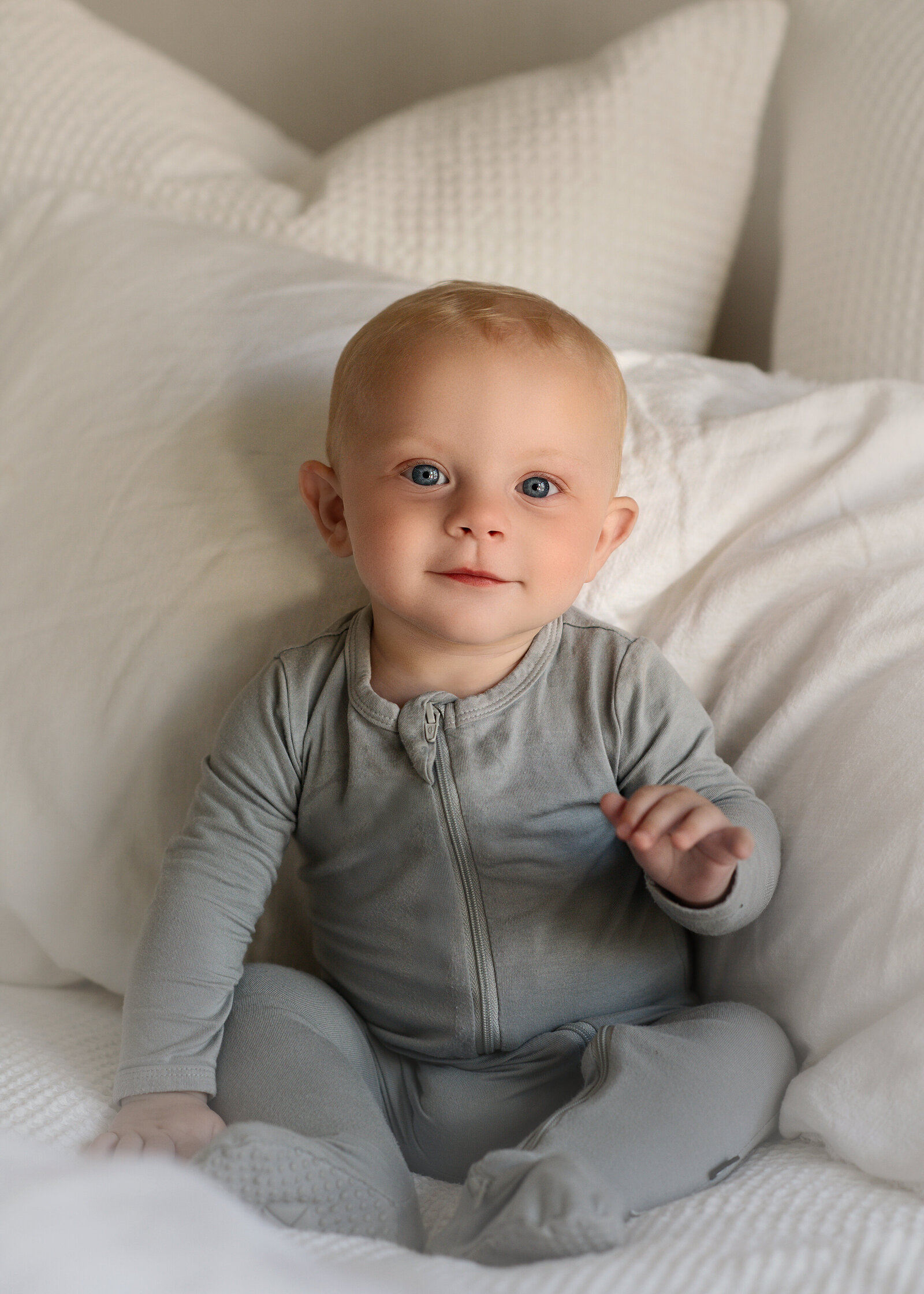 A baby with short, light blonde hair, dressed in a gray zip-up onesie, sits upright on a white bed while looking toward the camera with a relaxed expression
