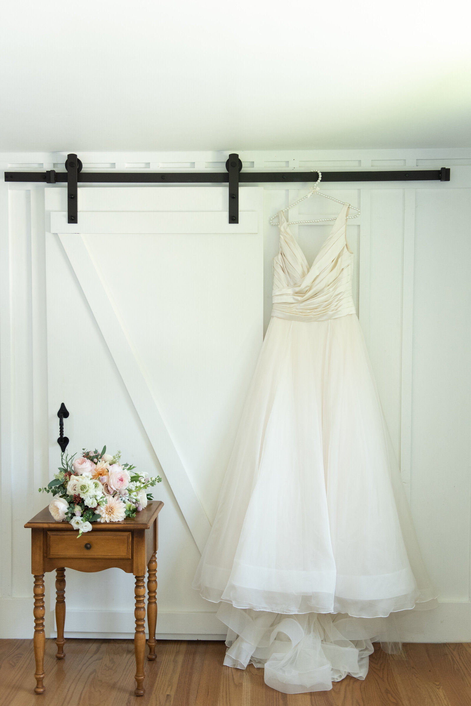 bridal dress hainging against white wall and brides bouquet on wood side table next to it