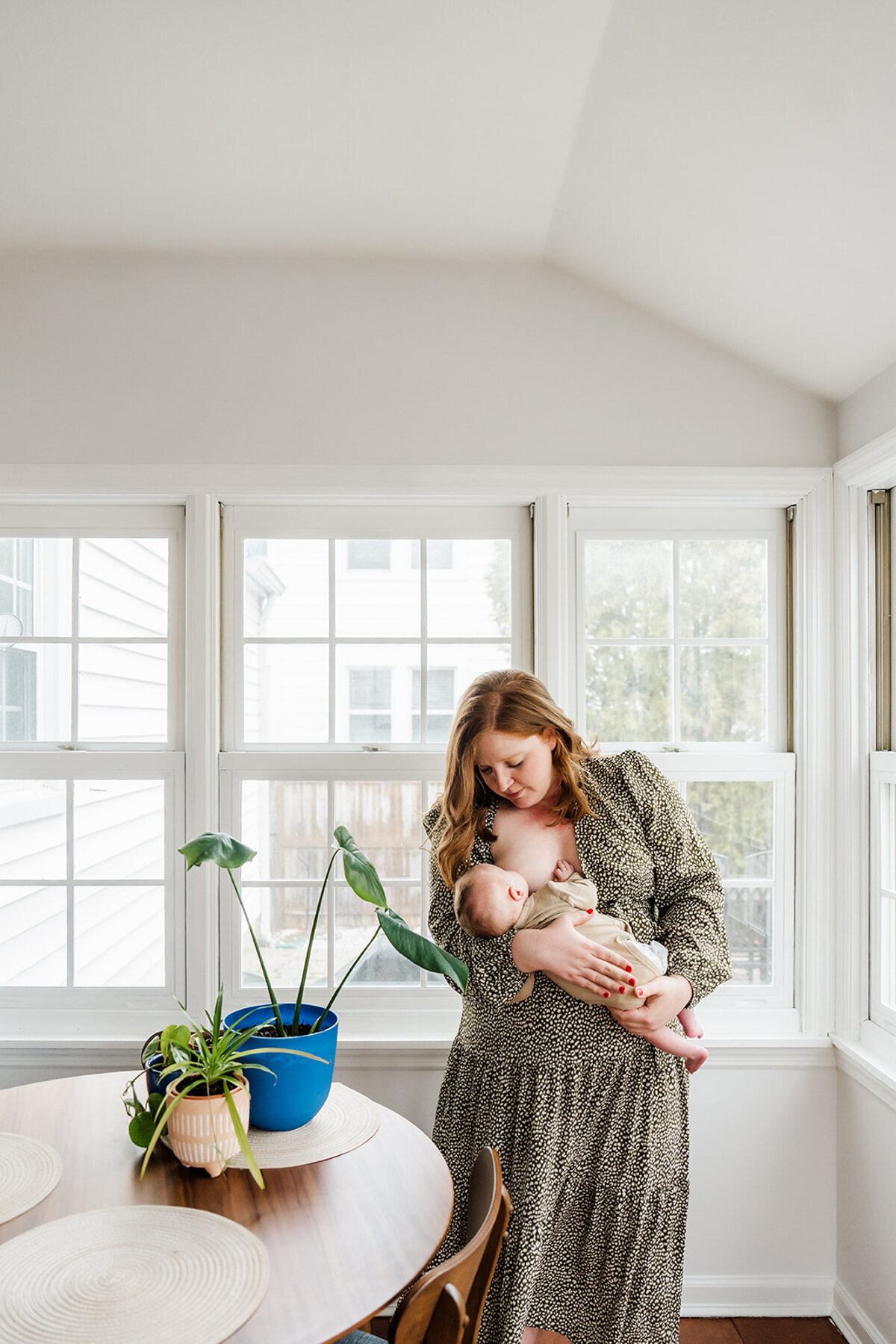 mother in green dress stands in a light filled kitchen nursing her baby boy