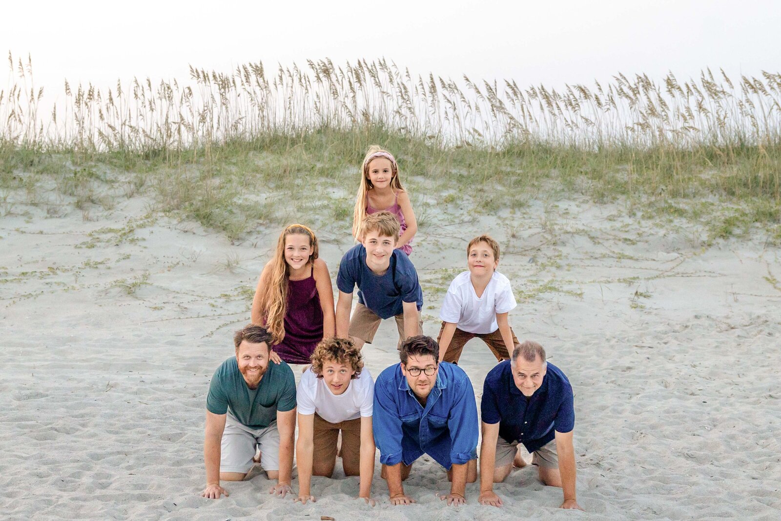 A family forms a playful pyramid on sandy dunes with sea oats swaying in the background. The joyful expressions and casual attire capture a fun moment by the beach, ideal for North Carolina family photography, showcasing lively family interactions.