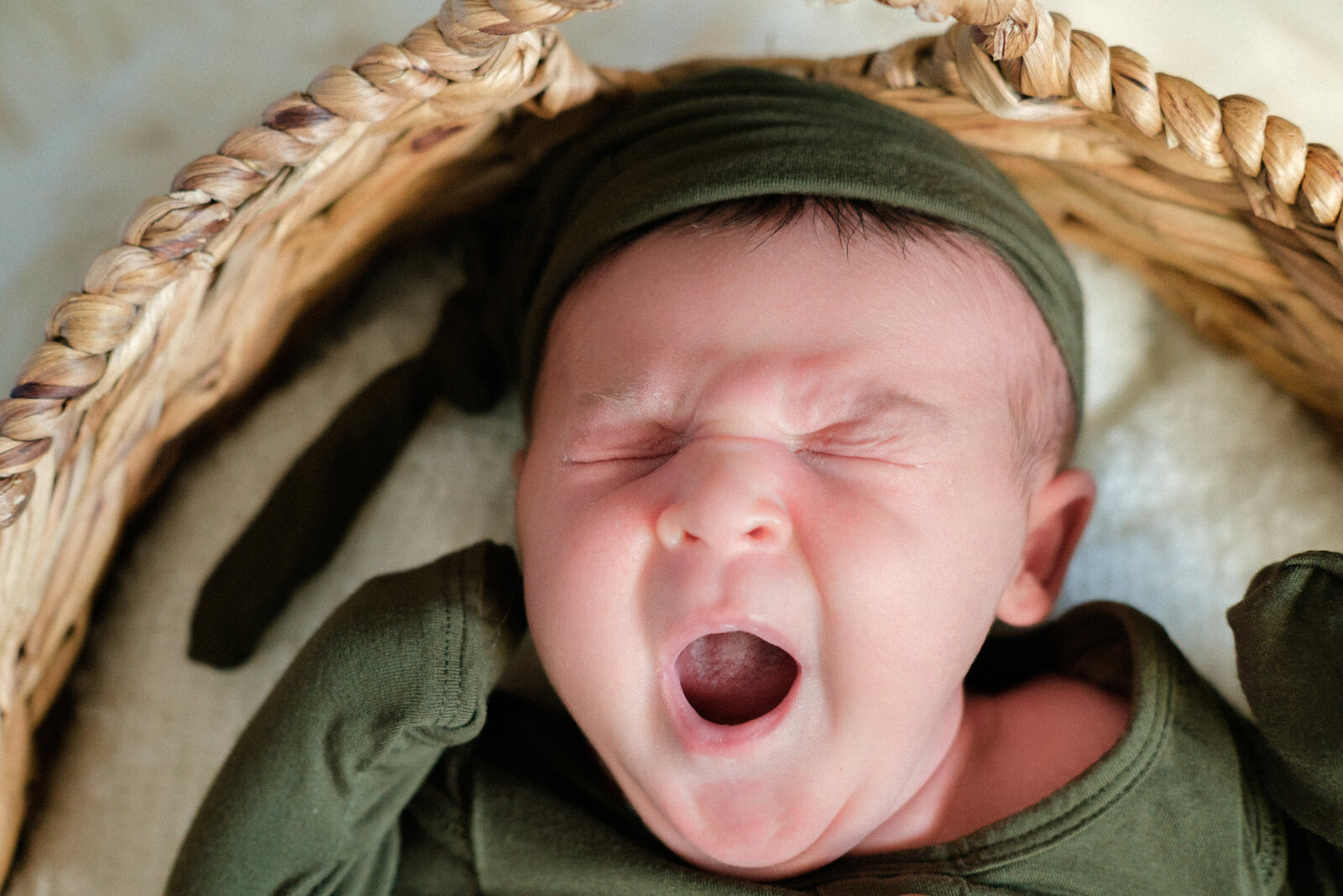 A baby boy yawns while laying in a basked and wearing a matching green hat and onesie