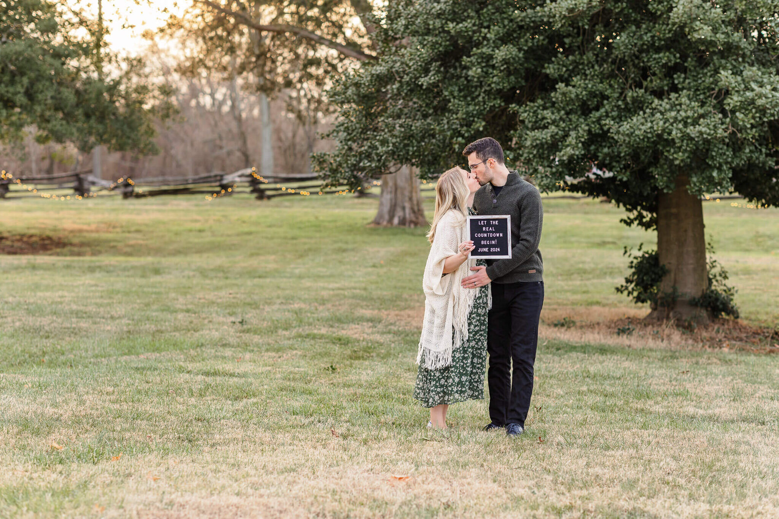 A mom and dad to be hugging with a sign announcing a pregnancy at a park at sunset.