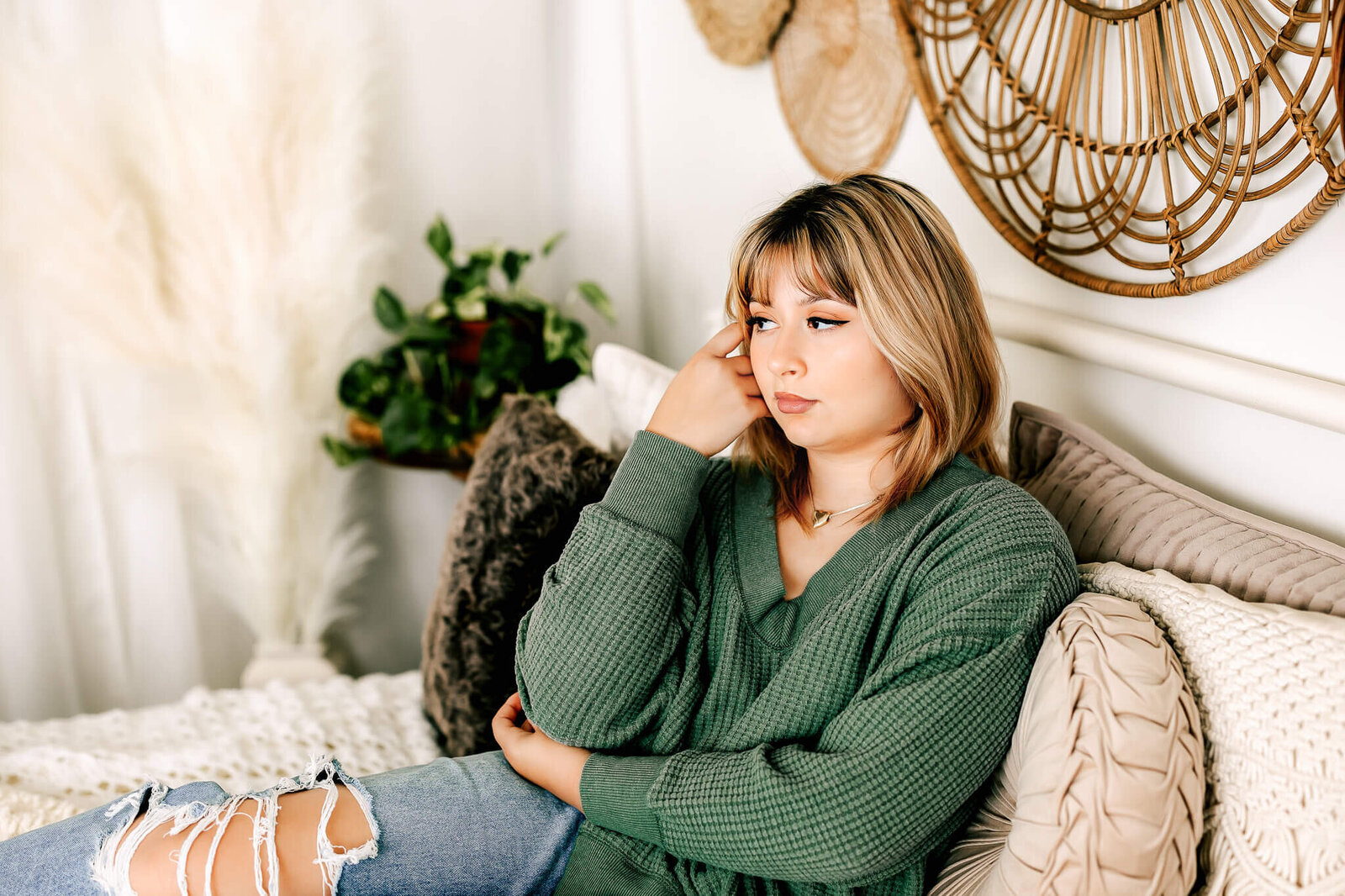 A person with shoulder-length hair sits thoughtfully on a bed with textured pillows. They're wearing a green sweater and ripped jeans. Decorative elements, such as a wicker wall hanging and a potted plant, are visible in the background.