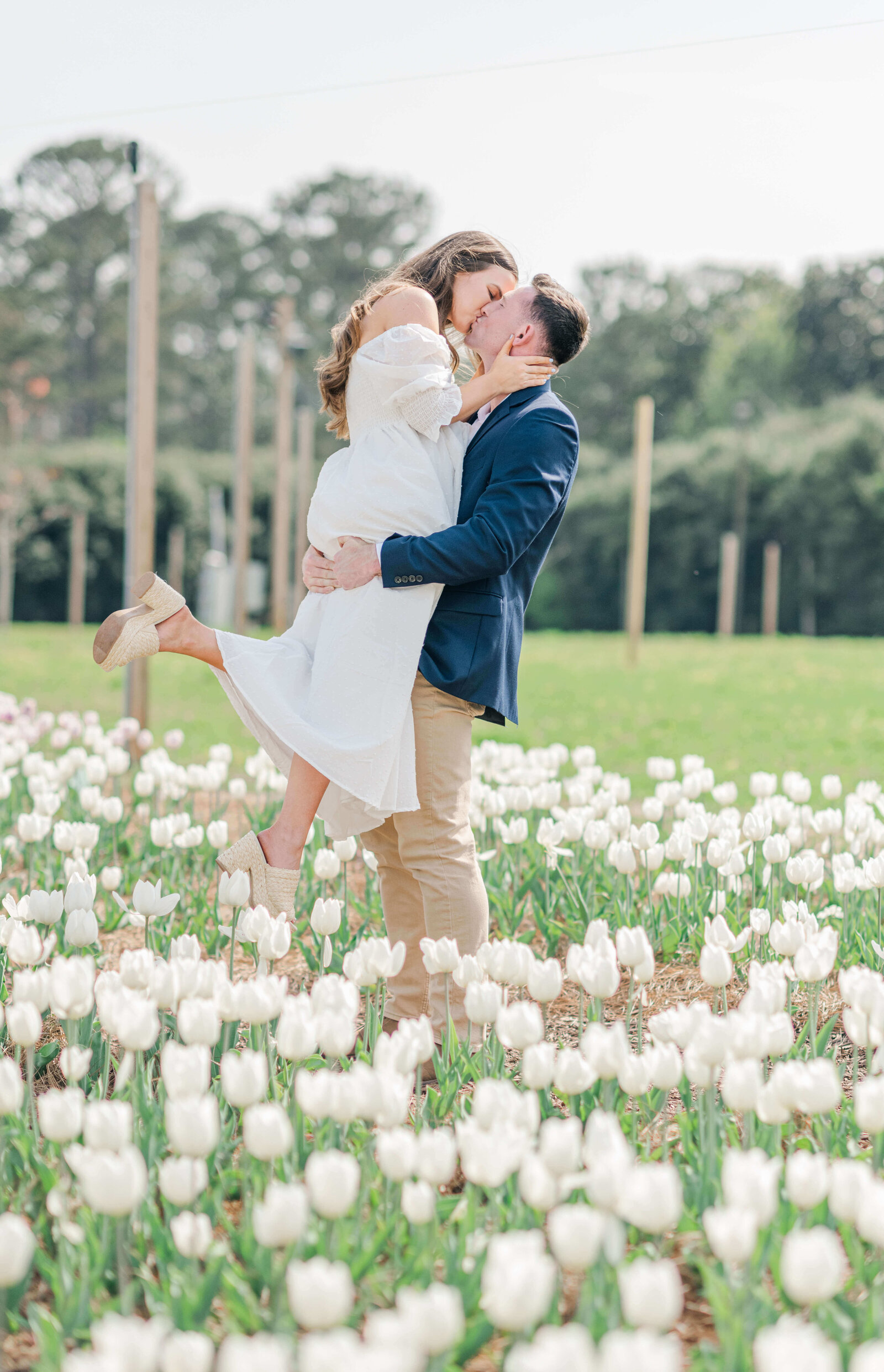 Engaged couple in a white tulip field with the groom holding up the bride for a kiss with a treeline background