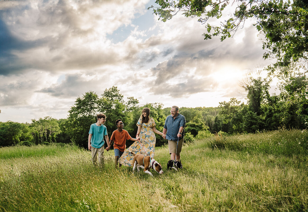 family walks holding hands in a breezy field with two sons and two dogs