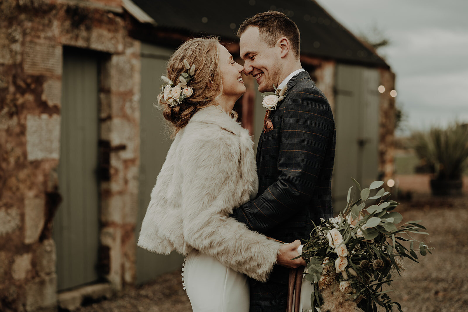 bride and groom hugging and smiling in front of outbuilding bride in fur jacket and holding bridal bouquet emotional aberdeen wedding photography