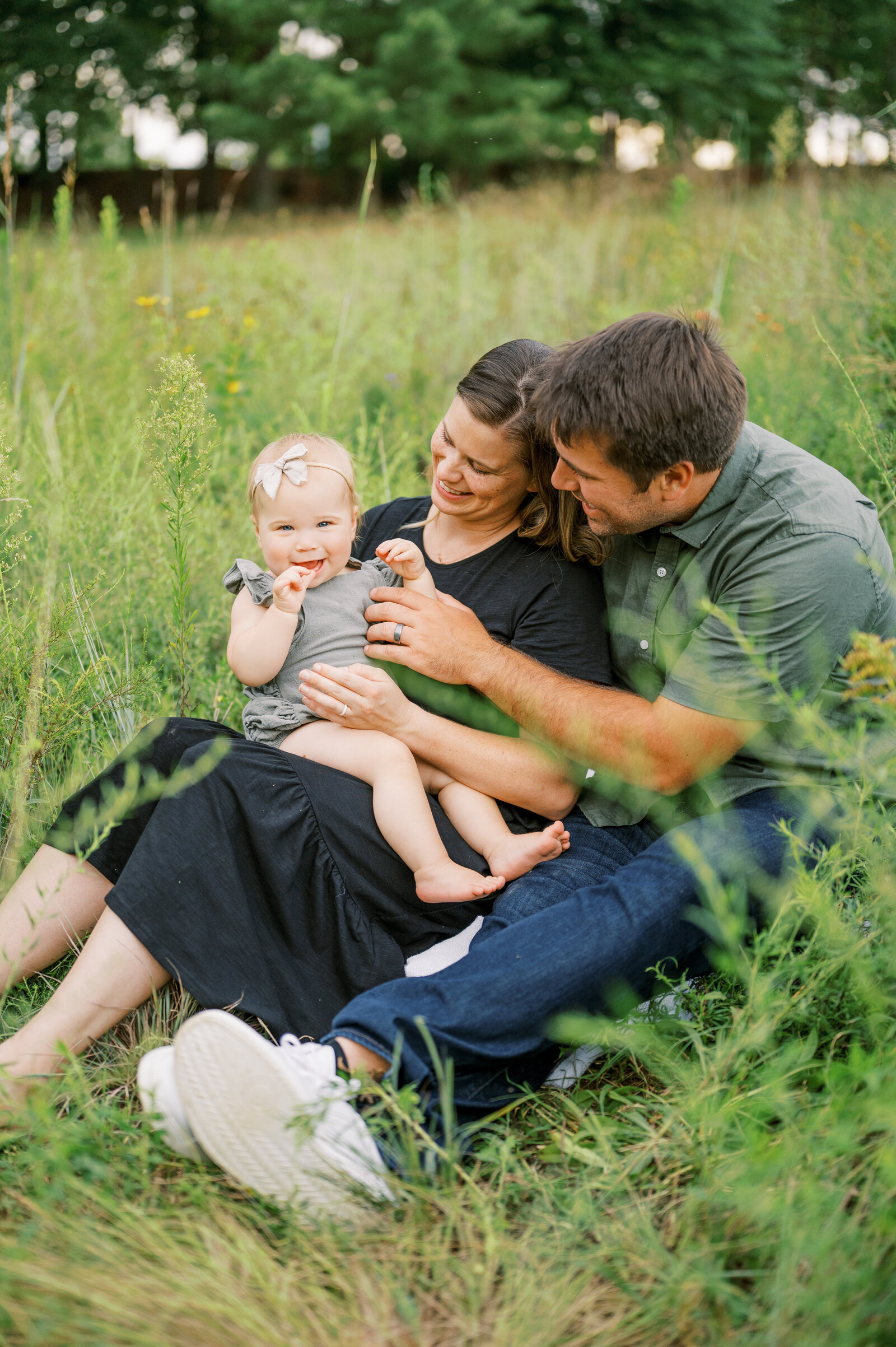 Mom and dad sit together in the grass holding one year old baby girl for family photo