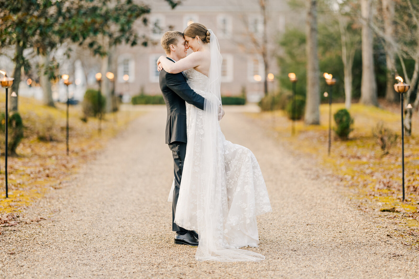 Groom lifts bride in a joyful celebration, her gown flowing as they laugh, capturing a playful and romantic moment.