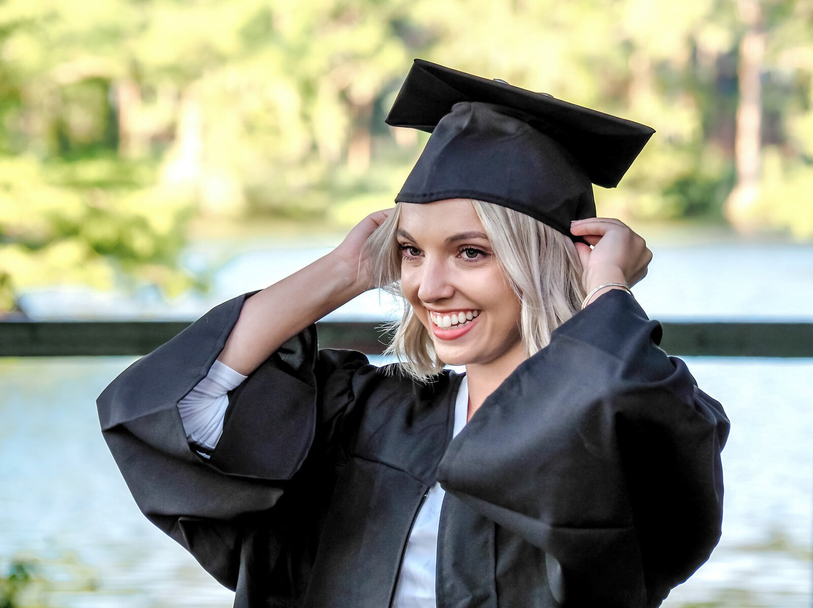 A smiling UNCW graduate in ablack cap and gown adjusting her tassel by a lakeside in Wilmingon, North Carolina. This joyful portrait captures the excitement of graduation, ideal for those seeking professional senior photography.