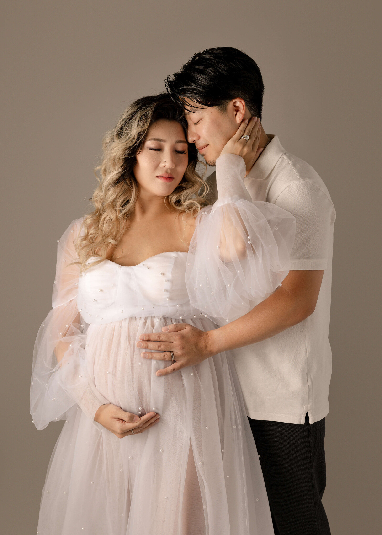 Husband and wife posed in studio with heads touching and eyes closed, momma is wearing white pearl dress by Ashley Nicole.