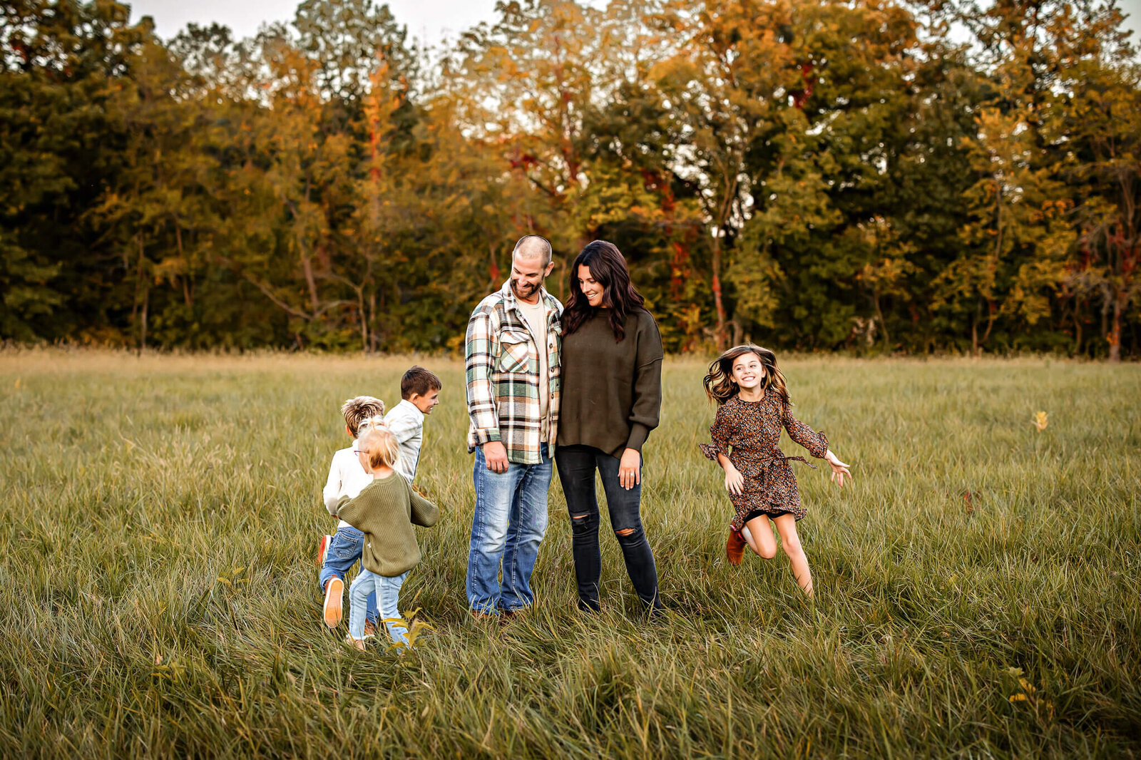 A family with two adults and four children stands in a grassy field surrounded by autumn trees. The children play and run around the smiling parents, capturing a moment of joy reminiscent of outdoor portraits taken by a skilled Bloomington family photographer.