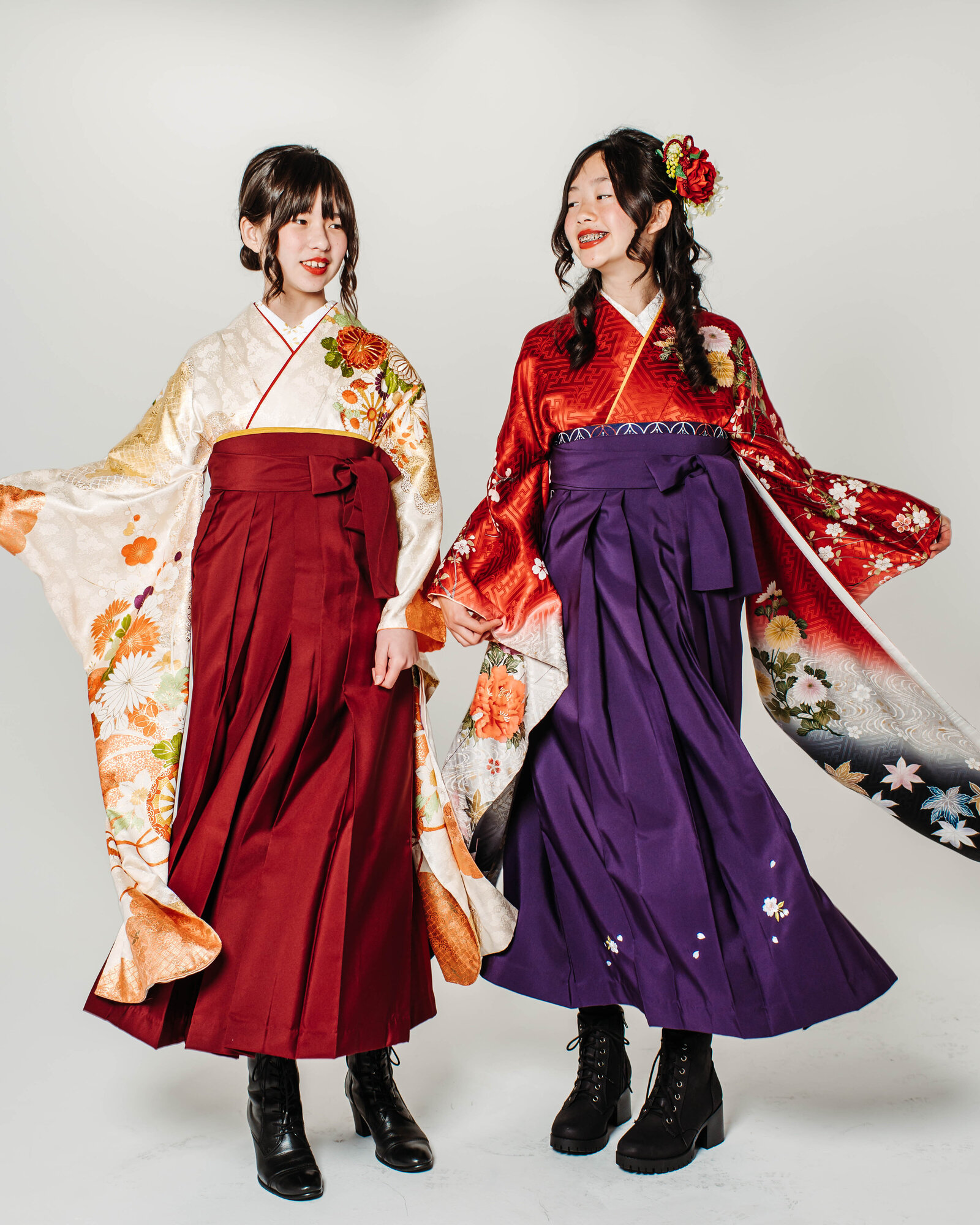 Two girls in hakama with floral kimono, one in red and one in purple, smiling together for a graduation photo, kimono rental Los Angeles. A beautiful look for LA graduation kimono and hakama rental | 卒業式 赤と紫の振袖袴を着た二人の女の子が一緒に笑顔を見せる ロサンゼルス着物レンタル