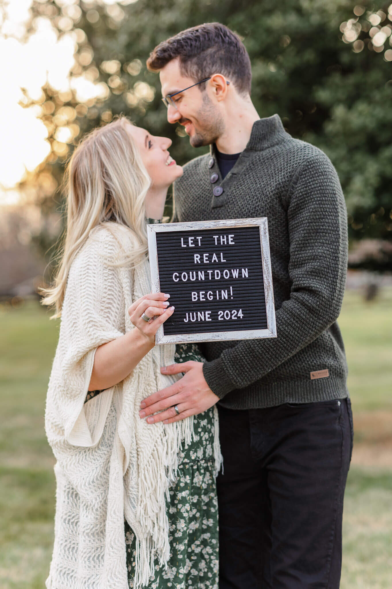 A couple looking into each other's eyes and holding a sign announcing their pregnancy at a NOVA park.