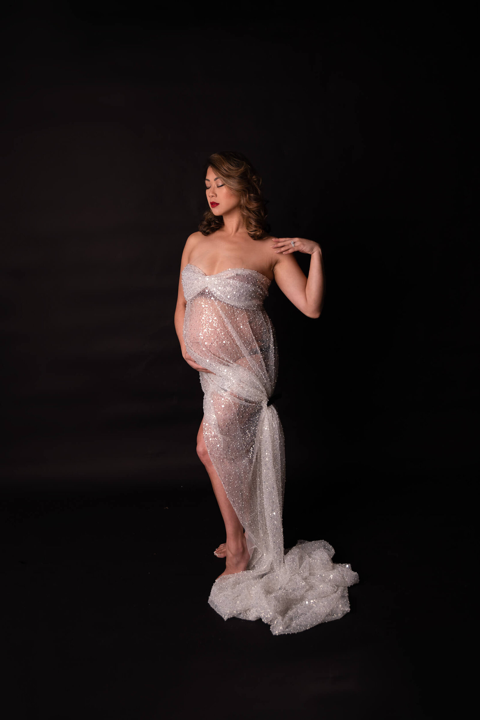 A mother-to-be stands in a shimmering silver sheer fabric gown against a dark backdrop, her poised stance and red lips enhancing the luxurious feel.