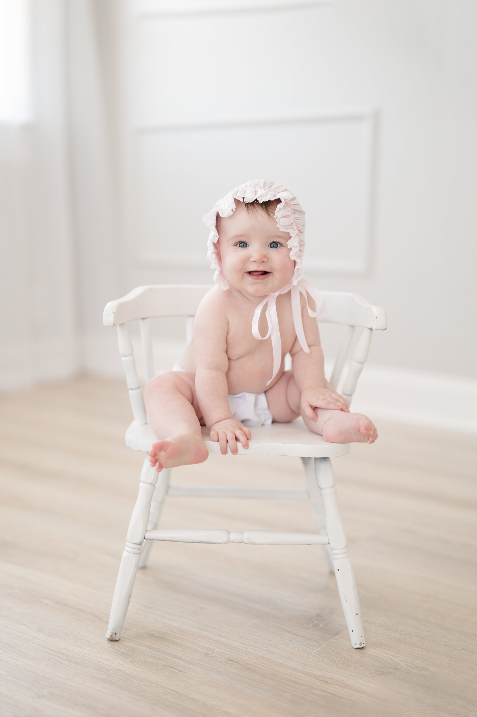 Baby girl wearing a bonnet and diaper cover while sitting on a white chair and smiling