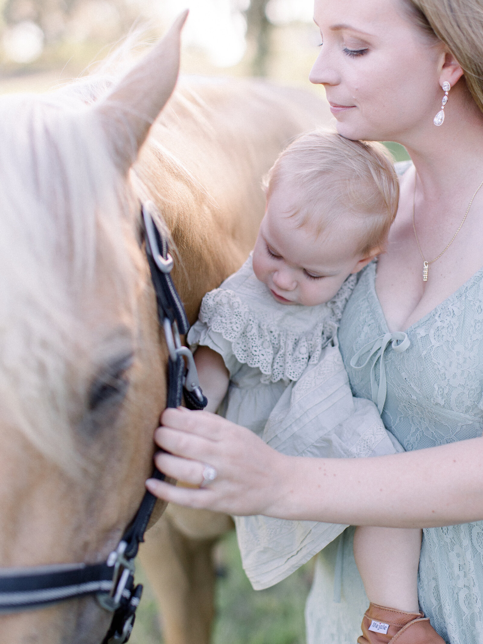 Closeup of mother in a sage lace gown holding her baby in a ruffled sage dress, petting their palomino horse on a horse farm in soft afternoon light by NH newborn photographer Fieldstone Studio. 