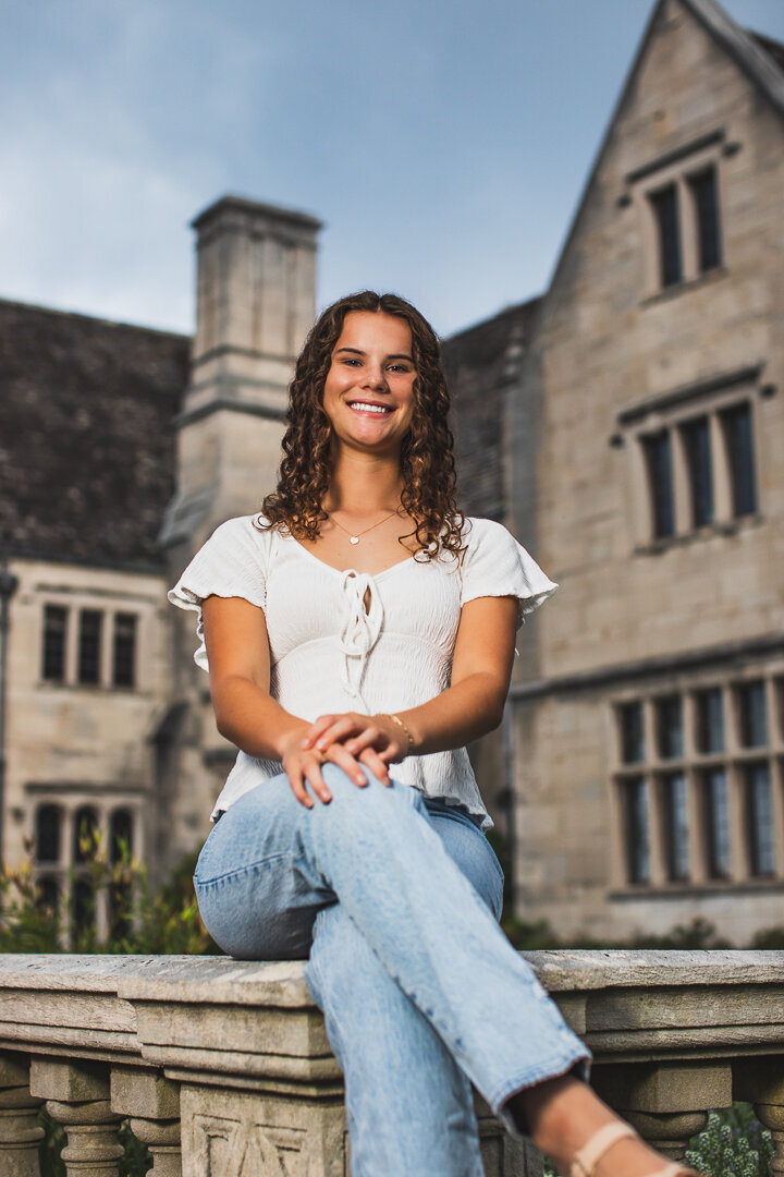 This curly haired senior girl poses with her hands on her knees a top the wall at Heartwood acres mansion.