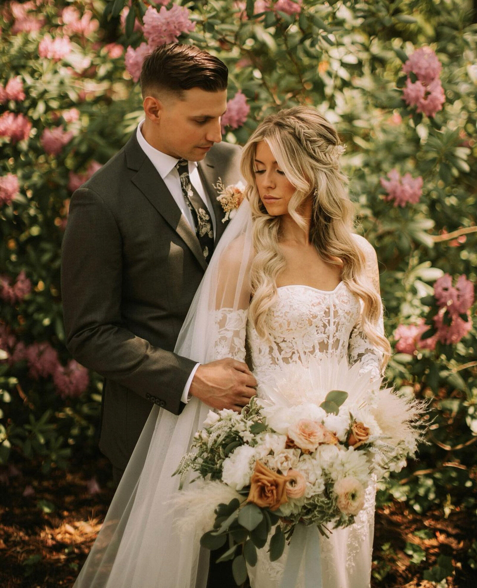 bride wears a lace wedding gown and holds a bouquet of white and peach flowers, while the groom, in a dark suit, gently places his hand on the bride's arm