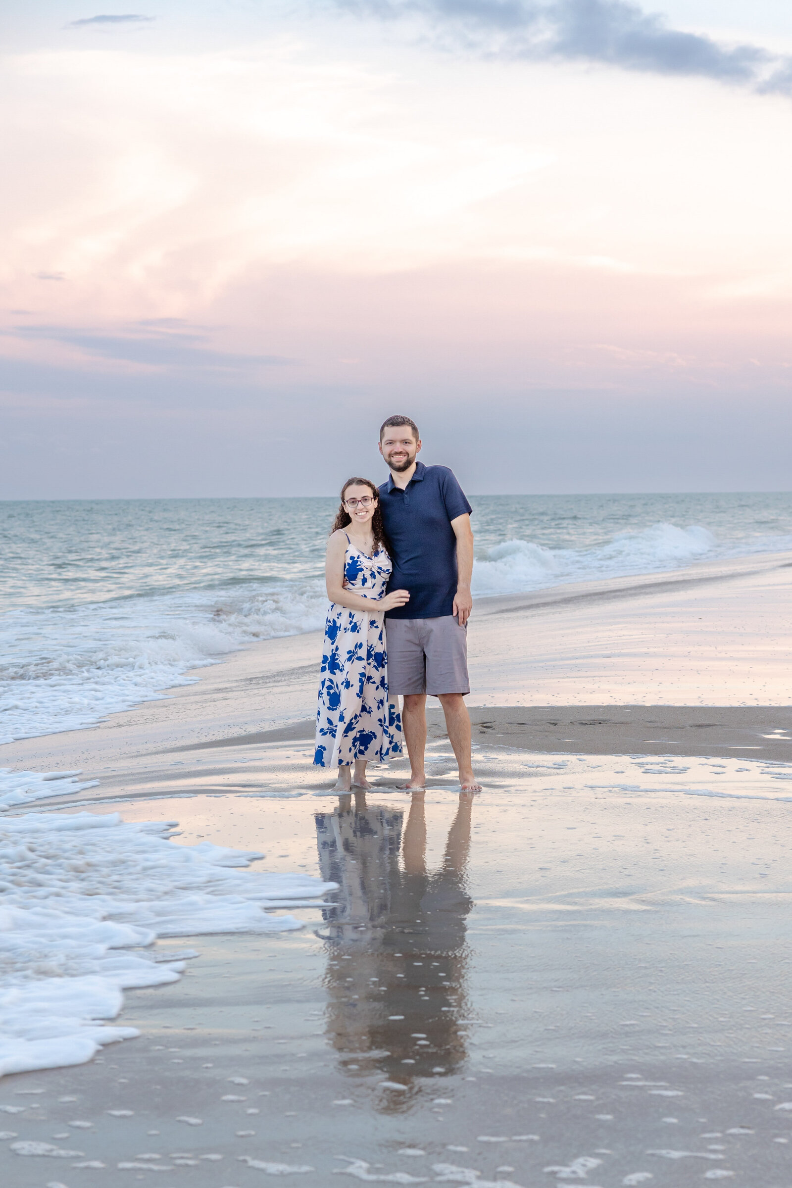A couple stands on a serene beach at sunset, their reflections mirrored in the wet sand. The woman wears a flowing floral dress, and the man sports a navy shirt and shorts. Gentle ocean waves and a colorful sky create a tranquil atmosphere, highlighting the romantic style of North Carolina engagement photography.  