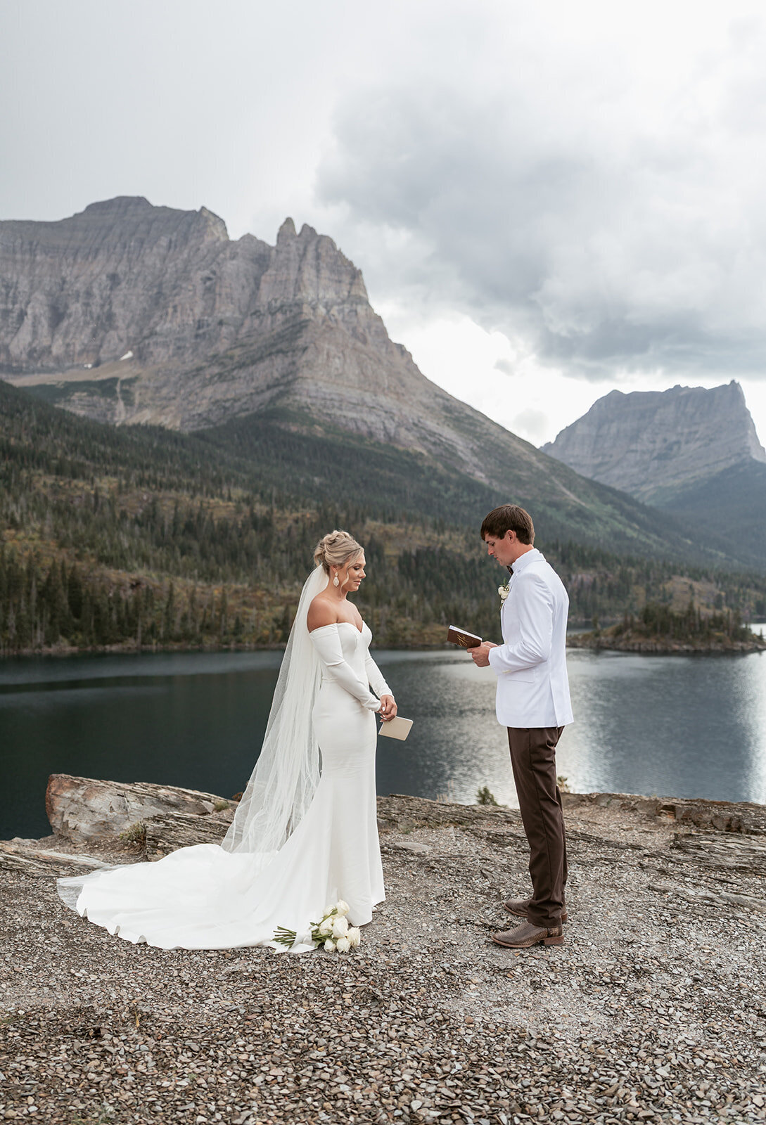 A bride and groom exchange vows beside a mountain lake in Glacier National Park, surrounded by dramatic peaks and overcast skies, captured by Sydney Breann Photography during their intimate Montana elopement.