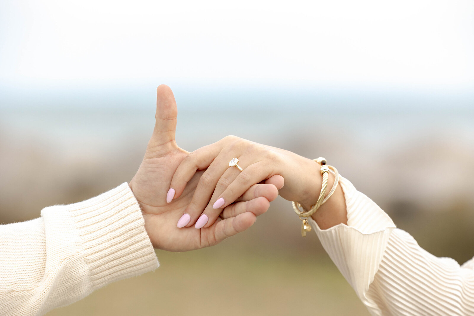 A close-up of a couple’s hands intertwined, showcasing a beautiful ring against a blurred beach backdrop. This image highlights the intimacy of engagement moments, reflecting the benefits of combined photography and videography services for weddings in NC.