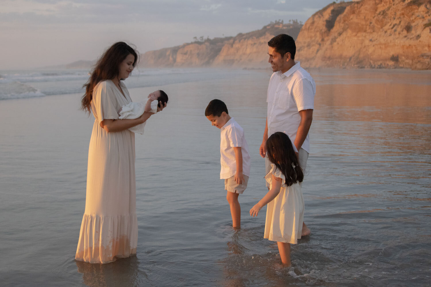 Family of four, standing by the water at a beach, with a mother holding their newborn