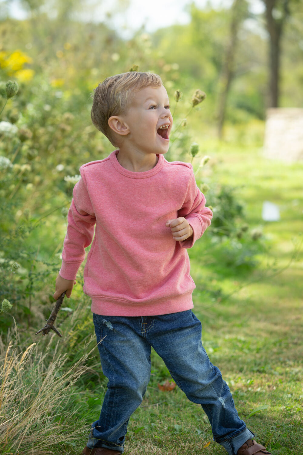 A young boy wearing a pink sweater and jeans is standing in the grass.