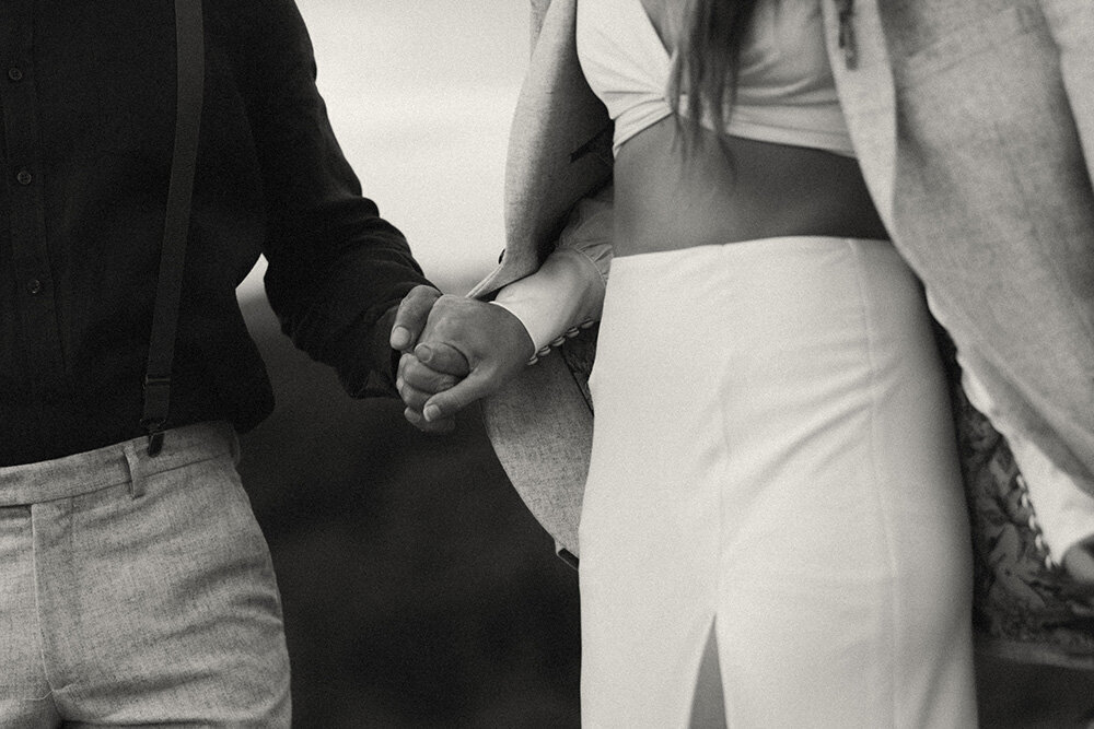 Black and white Close-up of an elopement couple holding hands, featuring modern wedding attire and candid connection, captured by elopement photographer.