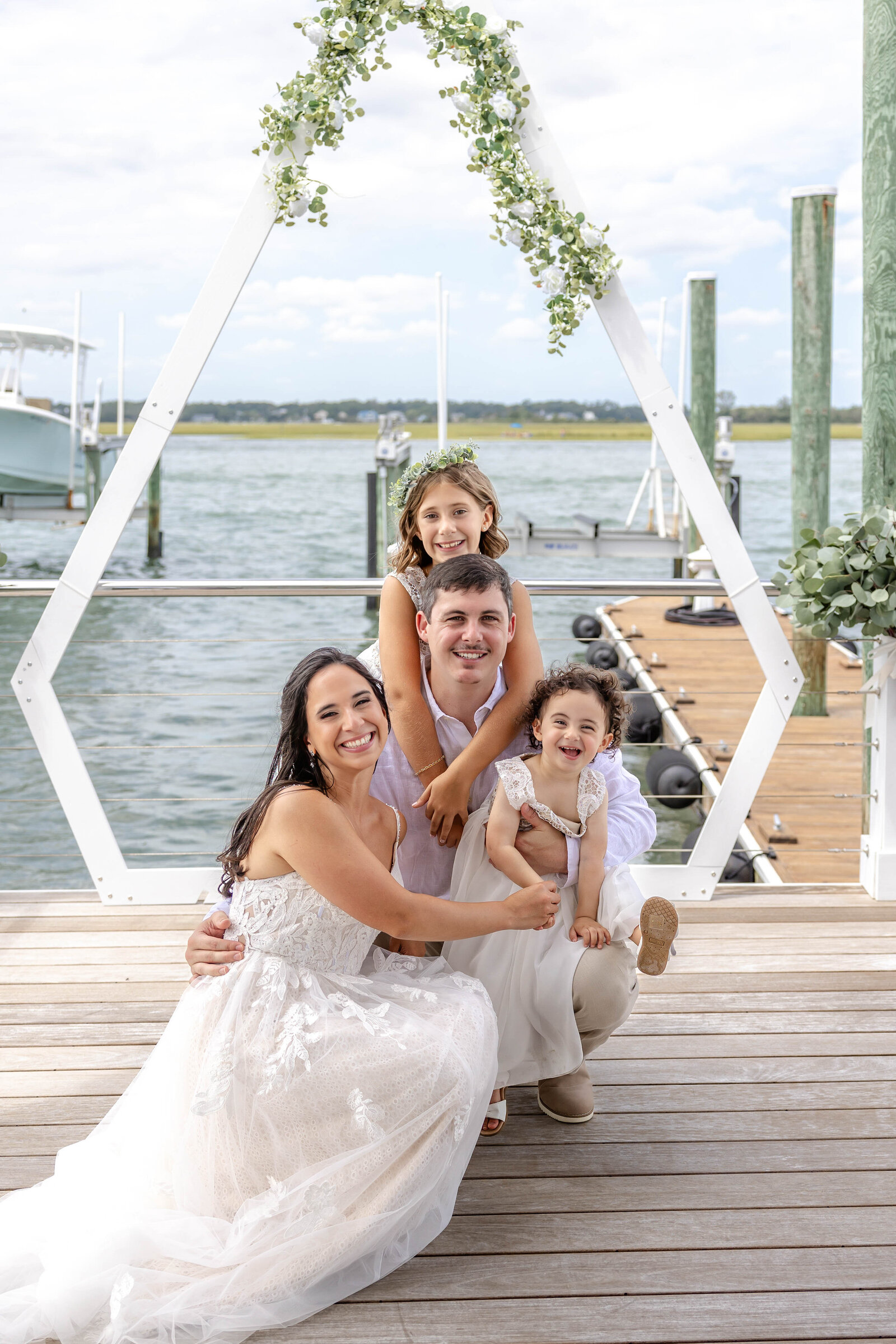 The bride and groom pose with two children under a geometric white wedding arbor adorned with greenery, captured on a dock during a Wrightsville Beach, NC wedding, blending family moments with coastal charm.