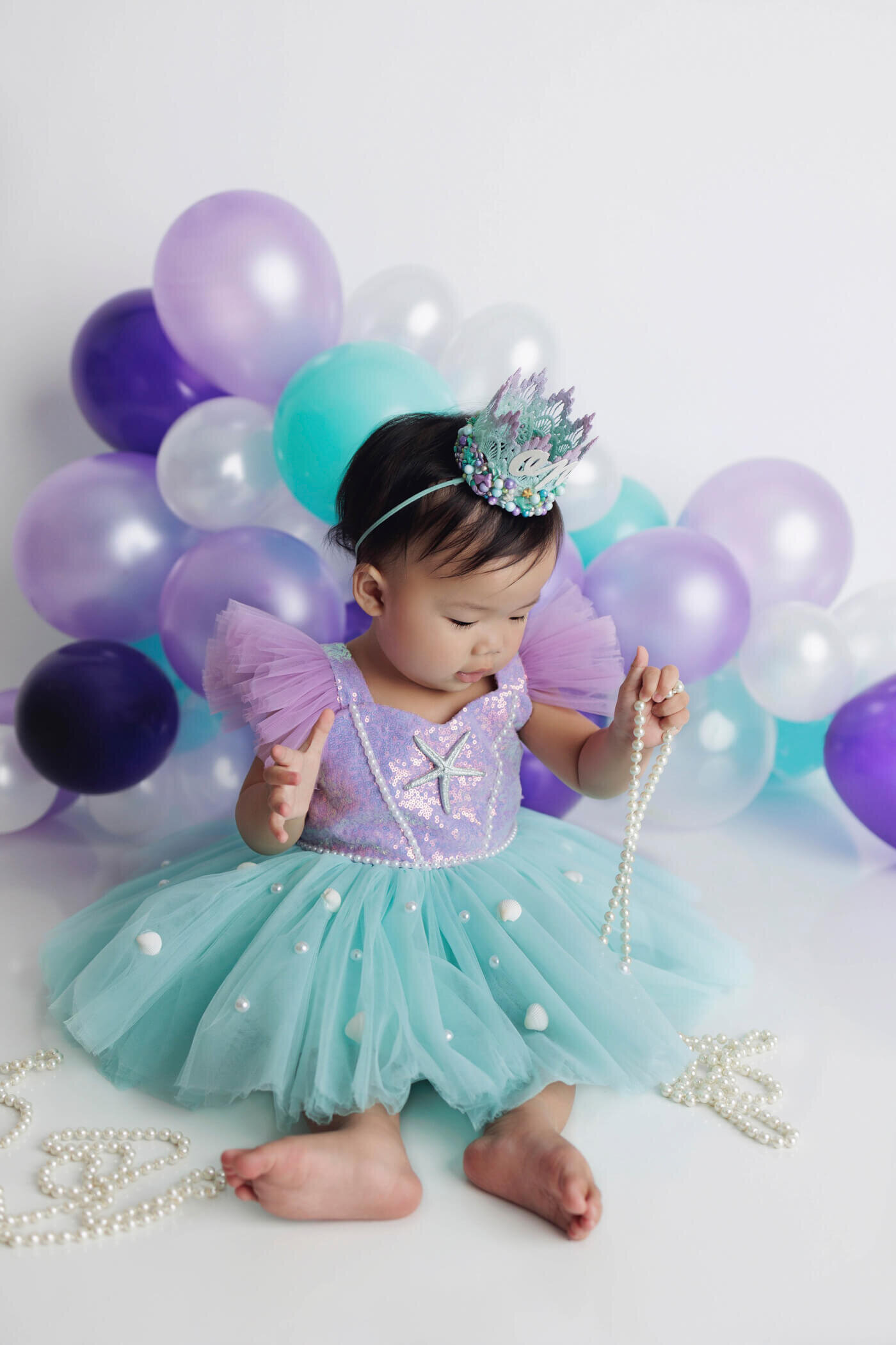 An adorable toddler in a princess-themed outfit sits on a white floor with a backdrop of purple, teal, and white balloons. The child wears a lavender and teal dress adorned with pearls and a starfish design, a matching crown, and holds a string of pearls while looking down, creating an enchanting and magical scene.