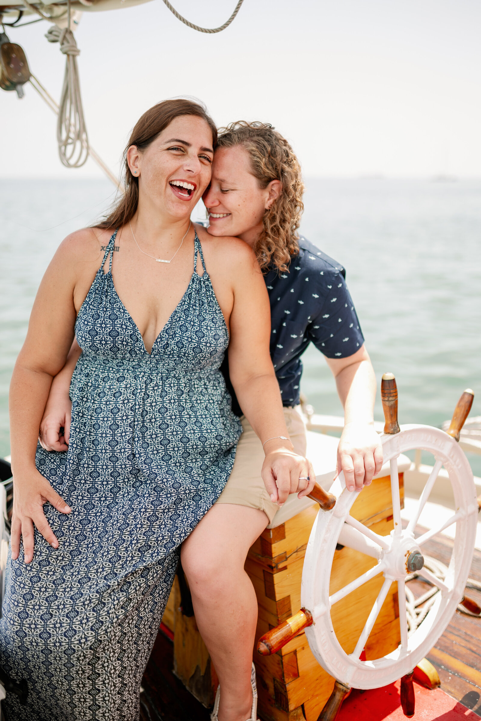 engaged lesbian couple sitting at the helm on a sailboat, laughing